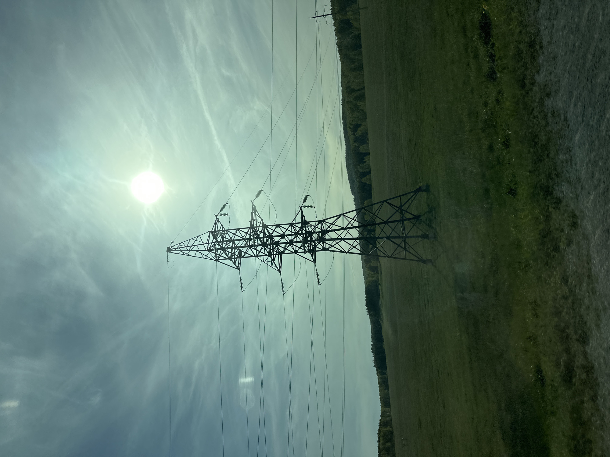 The image shows a high-voltage transmission tower in a grassy field under a partly cloudy sky with the sun visible. A line of trees is on the edge of the field. Other transmission lines extend into the distance. The location is Unknown, Unknown.