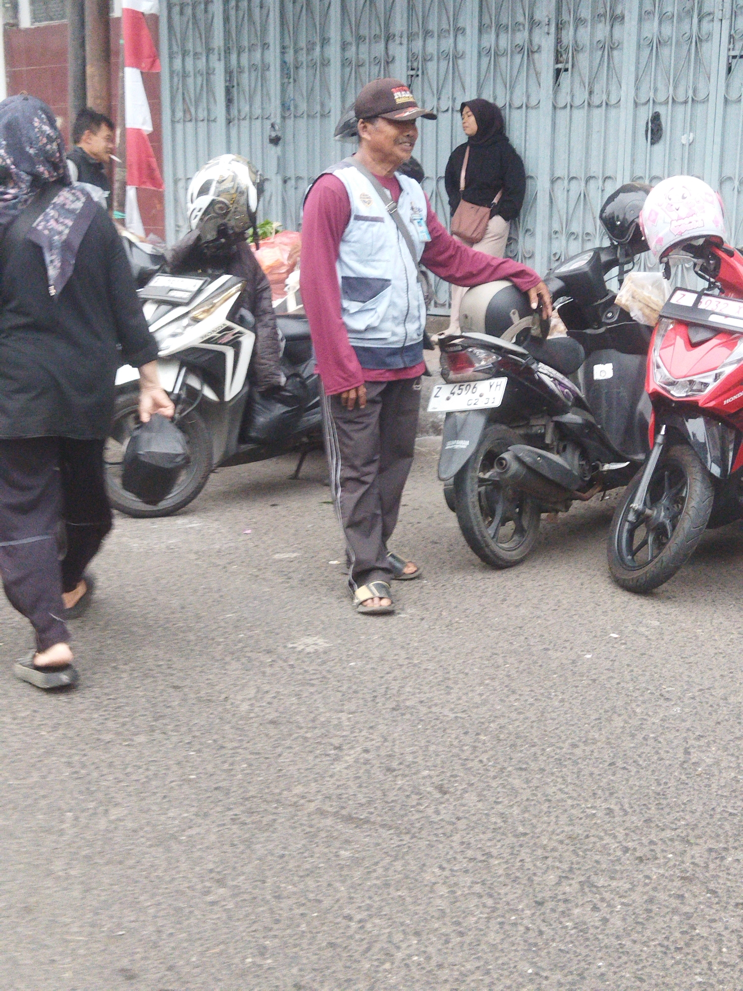 The image captures a street scene in Banjar, Indonesia, with several people and parked motorcycles. In the foreground, a man wearing a dark baseball cap, a blue vest over a maroon long-sleeved shirt, and dark pants with a white stripe, is standing next to a black motorcycle. He is looking to his right and appears to be talking to someone off-camera. His left hand rests on the motorcycle's seat. To his left, a woman wearing a dark hijab and black clothing is walking away from the camera, carrying a black bag in her left hand. Behind her, another motorcycle with a white and gold design is parked.
Further in the background, a woman in a black hijab and light-colored pants stands near a closed metal gate, with a pink and white helmet resting on a red motorcycle beside her. Another black motorcycle is positioned between the red one and the man in the blue vest. The ground is asphalt, and the overall environment suggests a public area, possibly a roadside or parking lot. The presence of multiple parked motorcycles indicates a common mode of transportation in the area. The backdrop features a textured metal gate and a partially visible red and white striped object, possibly a flag or banner, attached to a pole.