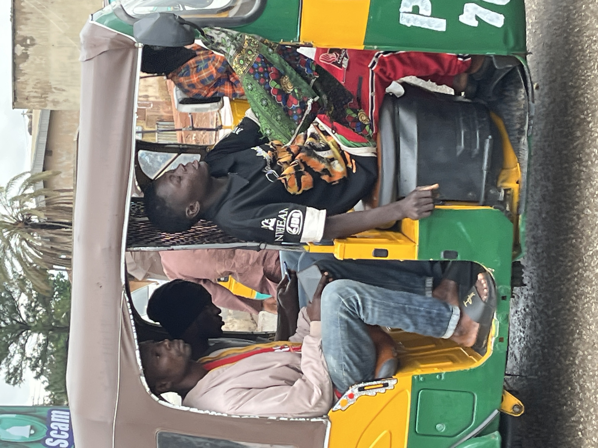 In Gura Top, Nigeria, an auto-rickshaw is stopped on an asphalt road. Inside, four individuals are seated. The driver, wearing a black t-shirt with a graphic design, is in the front passenger seat. To his left, a man in a light pink jacket and blue jeans is looking at his phone. Behind the driver, a person in an orange and blue plaid shirt is visible, and next to them, another person is wearing a red tracksuit with green and white accents. The auto-rickshaw is predominantly yellow and green, with some signs visible on its side. The background shows buildings and palm trees, suggesting an urban or semi-urban environment.