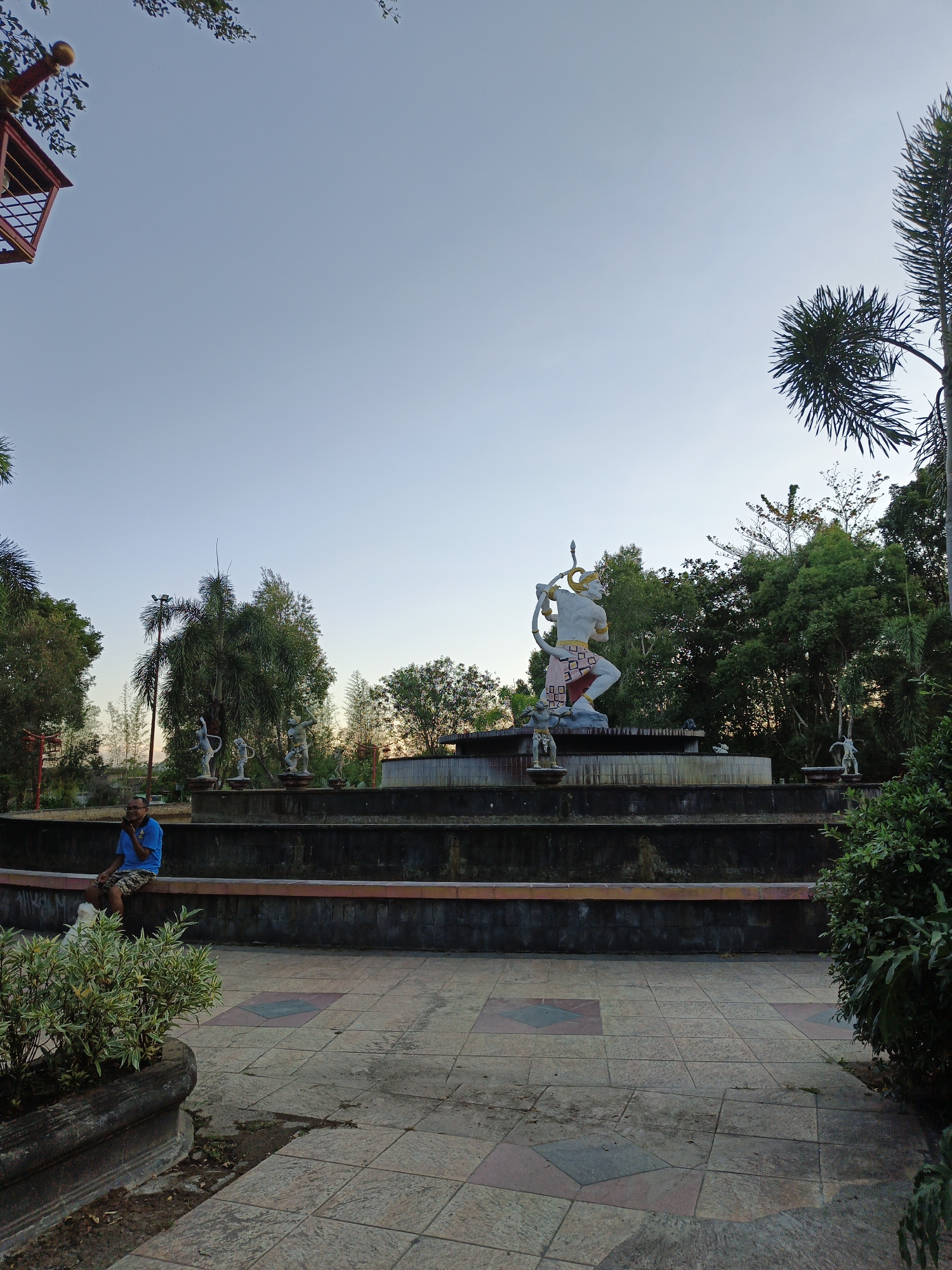 The image depicts a public park area with tiered water features and statues. In the foreground, a man wearing a blue t-shirt and patterned shorts sits on the edge of a fountain, looking at his phone. A white dog is visible near him. The background features a large statue of a multi-armed figure holding a bow and arrow, as well as several smaller statues of figures in dynamic poses. Lush green trees and vegetation surround the statues and water features. The sky is clear and appears to be either early morning or late afternoon. The paving in the foreground is tiled with a diamond pattern. This scene is located in Bareng Lor, Indonesia.