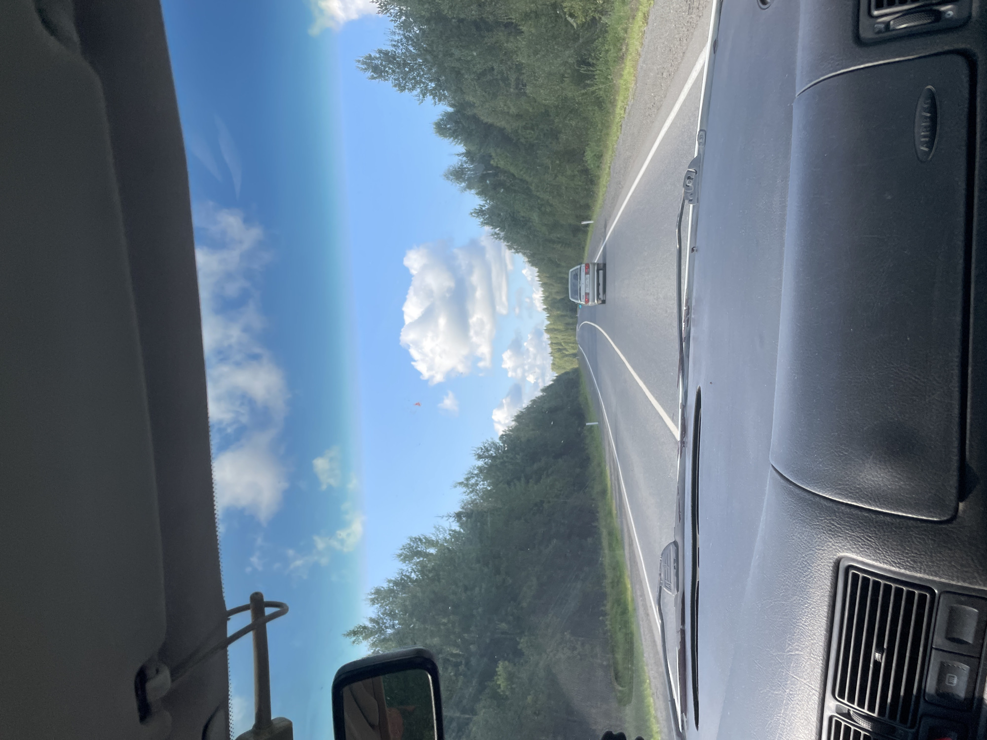 The image shows a view from inside a car traveling on a two-lane road through a forest. The sky is blue with scattered white clouds. The road is paved with white lane markings and has a paved shoulder. Trees line both sides of the road. In the distance, another car is visible. The interior of the car, including the dashboard, steering wheel, and windshield, is partially visible in the foreground.