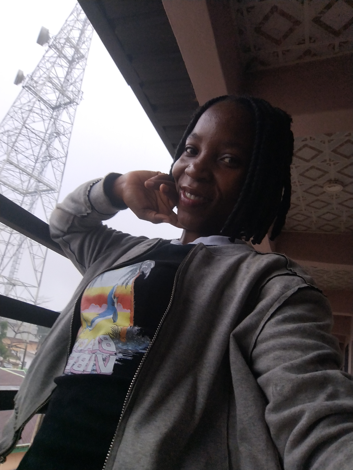 In a selfie taken from a slightly upward angle, a young Black woman with braided hair and a gray zip-up jacket over a graphic t-shirt smiles at the camera. She rests her right hand against her cheek, with her left hand near her chest. The background features a large, lattice-like communication tower against a hazy sky, suggesting an outdoor urban setting. A portion of a building with decorative tiling is visible above and behind her. The image appears to have been captured in the city of Iguosa, Nigeria.