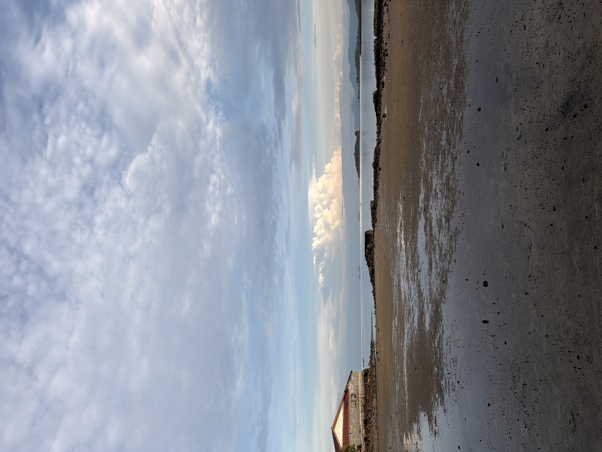 A wide-angle vertical photograph captures a serene coastal scene at low tide in Koh Samui District, Thailand. The foreground is a wet, sandy beach dotted with small tide pools that reflect the sky. Dark, textured sand stretches across the bottom of the frame. In the middle ground, a rocky shoreline meets a calm body of water. To the left, an old, weathered building with a red-tiled roof and brick walls stands precariously close to the water's edge. The background features a calm sea meeting a hazy horizon with distant, silhouetted islands and mountains. The sky above is a dramatic canvas of soft blues and whites, filled with scattered clouds that range from wispy cirrus to large, puffy cumulus formations, illuminated by soft, diffused light, suggesting it might be early morning or late afternoon. There are no people visible in the image.