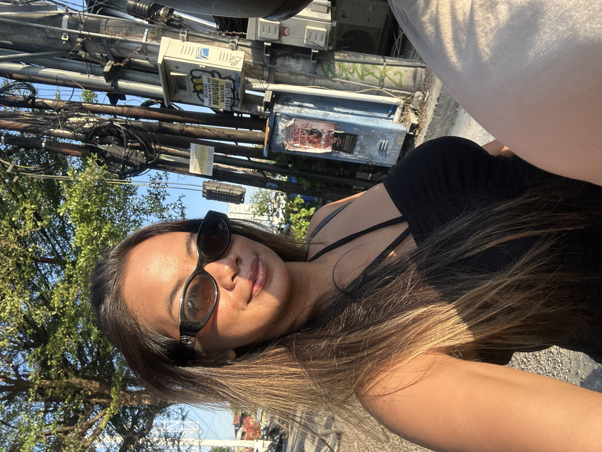 This is a vertical, low-angle, outdoor selfie taken on a sunny day in North Kuta, Indonesia. The subject is a young woman with long, highlighted brown hair, wearing large, dark-rimmed sunglasses and a black tank top. She is looking at the camera with a soft smile. The background is dominated by a tangle of electrical wires and poles, with some green trees and a bright blue sky visible. There are several metal boxes attached to the poles, some with signs. One sign, visible in the cropped images, has Indonesian text that appears to be an advertisement for land sales. Another sign reads "DIJUAL TANAH KAVLING". There are also posters on one of the boxes. The lighting suggests it is either late afternoon or early morning, with the sun casting warm highlights on the woman's hair and skin and creating shadows on the ground. The overall impression is one of a casual, everyday moment captured in a busy urban environment.