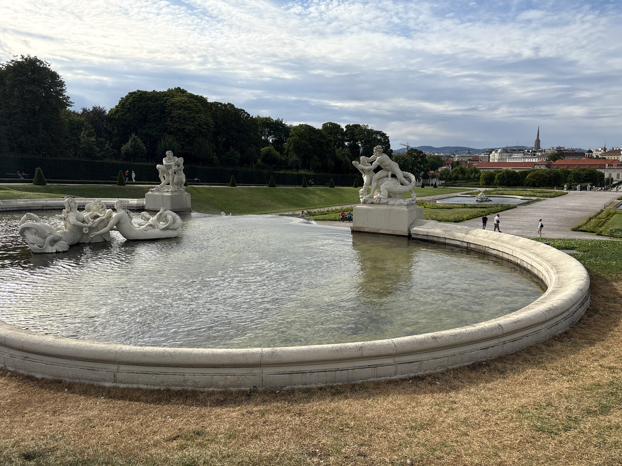 The image shows a fountain in the Belvedere Palace gardens in Vienna, Austria. The fountain features several white marble sculptures of mythological figures, including mermaids and a man wrestling with a dragon or sea creature. The fountain is circular with a wide, stone rim, and the water is clear, reflecting the cloudy sky above. In the background, there are manicured green lawns, trimmed hedges, and mature trees. Beyond the gardens, the cityscape of Vienna is visible, with buildings and a prominent church spire. Several people are walking in the gardens and near the fountain. The grass surrounding the fountain is dry and brown, suggesting a warm weather season.