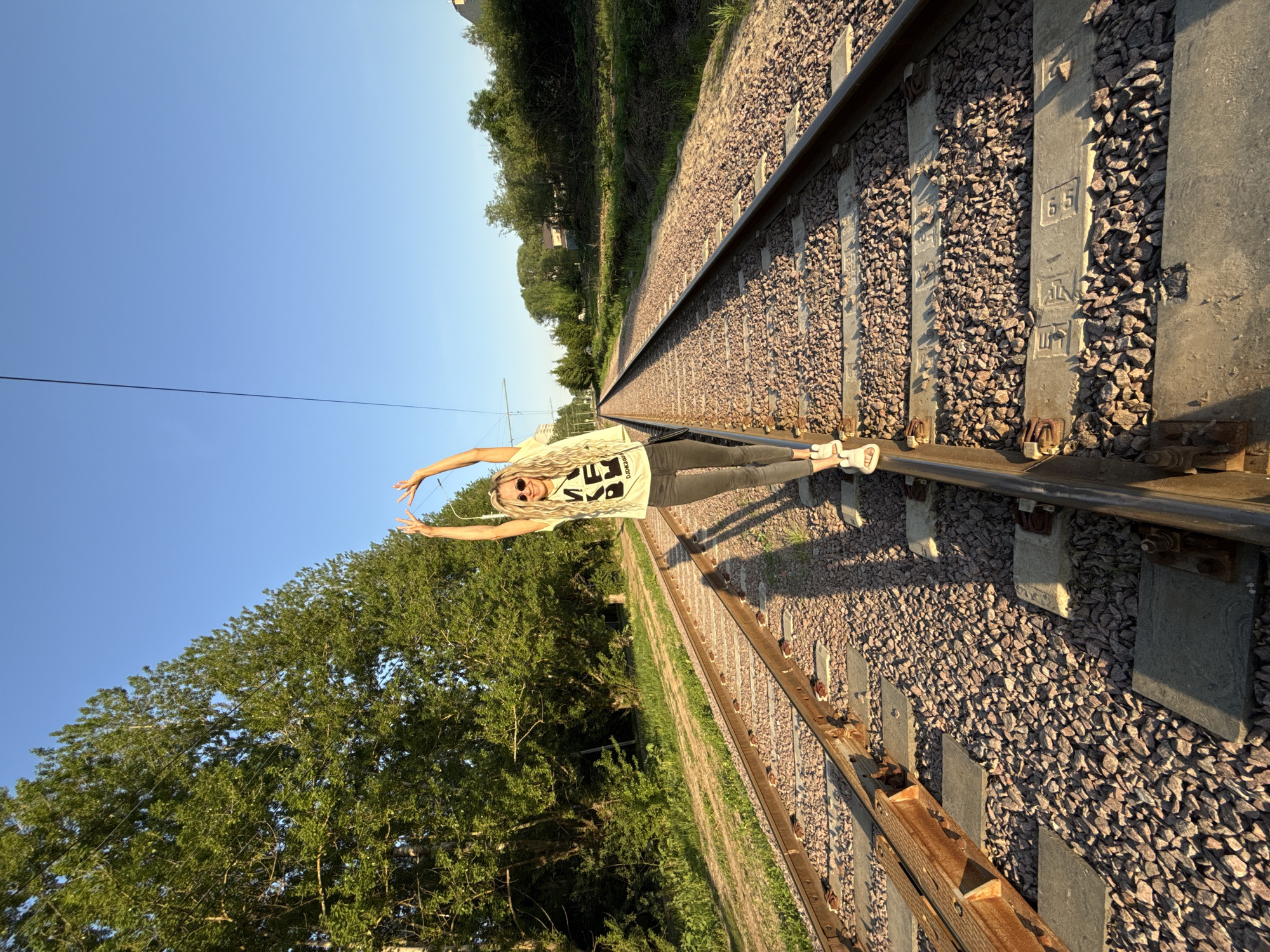 The image shows a woman standing on railroad tracks, likely an outdoor location near trees. She is wearing a T-shirt, leggings, and sandals. She is holding her hands above her head in a heart shape. The location is not confirmed to be Rusanovo, Russia and there is no evidence to verify this claim.