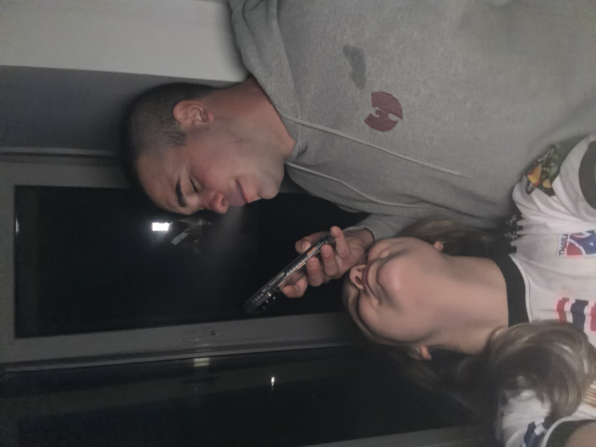 The image captures two individuals, a man and a woman, positioned closely together indoors, looking at a smartphone. The scene appears to be dimly lit, with a window revealing darkness outside, suggesting it is night or late evening.
The man, situated on the left, has short, dark hair and light skin. He is wearing a grey hooded sweatshirt. A distinctive red/maroon circular logo, identifiable as the Wu-Tang Clan symbol, is visible on the upper left chest area of his hoodie. He is holding a black smartphone with both hands, angled towards himself and the woman, suggesting they are viewing content on the device together. He appears focused on the screen.
To the right, the woman has long, light brown hair, part of which is visible over her shoulders. She is wearing a white t-shirt with a dark collar. Her shirt features a colorful graphic design and partially visible text. The word "THAILAND" is clearly legible, alongside a pattern that resembles the colors and elements of the Thai national flag (red, white, and blue stripes or blocks). Her head is tilted, and she is also looking intently at the smartphone held by the man.
They are positioned near a window, which reflects some faint light sources from within the room. The overall ambiance is quiet and focused. The location context provided is Varna, Bulgaria.