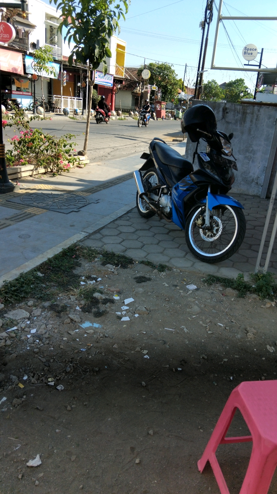 The image depicts a street scene in Batang, Indonesia. In the foreground, a blue and black motorcycle is parked on a hexagonal paved sidewalk. A black helmet rests on the motorcycle's seat. Behind the motorcycle, a concrete wall and a few scraggly plants are visible. The street, a manhole cover, and more hexagonal paving are also present. In the background, several businesses line the street, including one with a sign reading "Space Blue." A few motorcycles are seen in motion on the street, with riders wearing helmets. The sky is clear and blue. The overall impression is of a typical urban street environment.