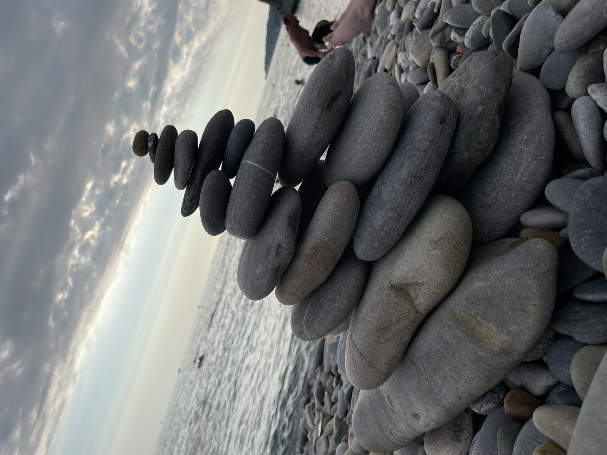 An outdoor, eye-level, medium shot captures a stack of large, smooth, gray river stones artfully balanced on a pebble beach. The stones are arranged in a tapering tower, with the largest at the bottom and progressively smaller stones stacked towards the top, culminating in a single, small, reddish-brown stone. The background features the calm, gray-blue water of the sea, with a few distant people swimming or wading. The sky above is overcast with dramatic, dark gray clouds streaked with lighter gray and white. The overall lighting suggests it is either early morning or late evening. The presence of a pebble beach indicates the location is a coastal area, consistent with Sochi, Russia, known for its pebble beaches along the Black Sea.