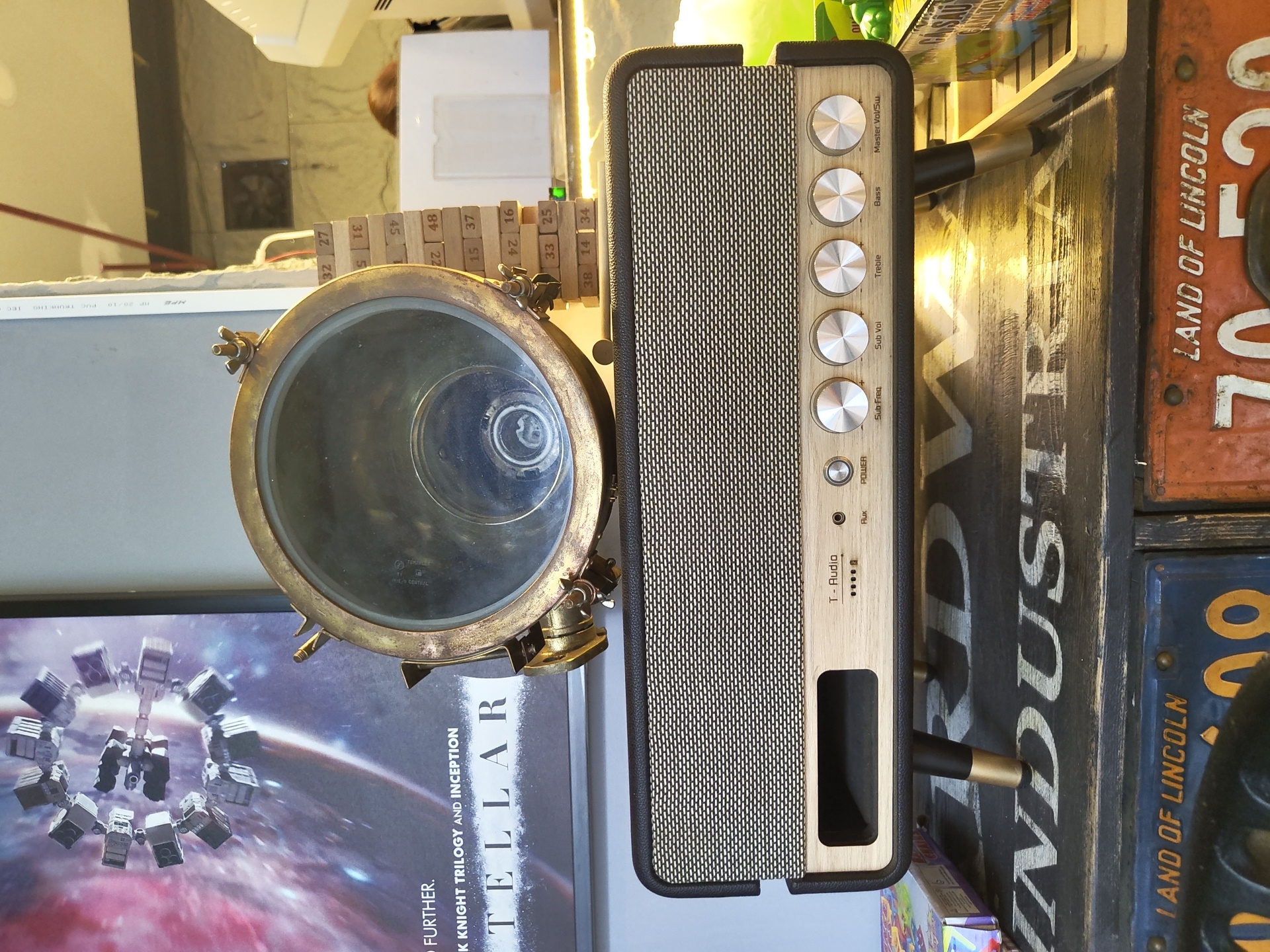 The image shows a close-up view of a vintage-style speaker with a wooden and black textured finish, mounted on a dark wooden surface with the word "INDUSTRY" painted on it. The speaker has a patterned fabric grille and several chrome knobs labeled "POWER," "Sub Freq," "Sub Vol," "Treble," "Bass," and "Master Vol/SW." To the left of the speaker, there's a poster for the movie "Interstellar." On top of the speaker, a brass fixture with a glass lens is visible. A stack of wooden blocks, possibly from a Jenga game, is positioned behind the brass fixture. The background includes parts of a wall, a white object, and a blurry figure of a person with brown hair. The lighting appears to be warm and slightly reflective. The scene is likely an indoor setting, possibly a shop or a home, and does not provide any specific indicators to determine its location in Nha Trang, Vietnam.