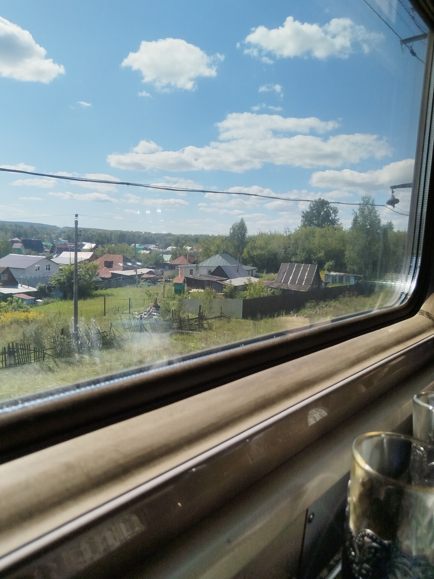 The image shows a view from inside a train carriage looking out of the window at a rural landscape. The scene depicts a village with houses, trees, and fields under a blue sky with white clouds. A power line runs horizontally across the sky. In the foreground, the interior of the train carriage is visible, including the window frame and two glasses on a table. The village appears to be located in Yumatovo, Russia, judging by the style of the houses and the surrounding vegetation. The time of day appears to be daytime, with bright sunlight illuminating the scene.