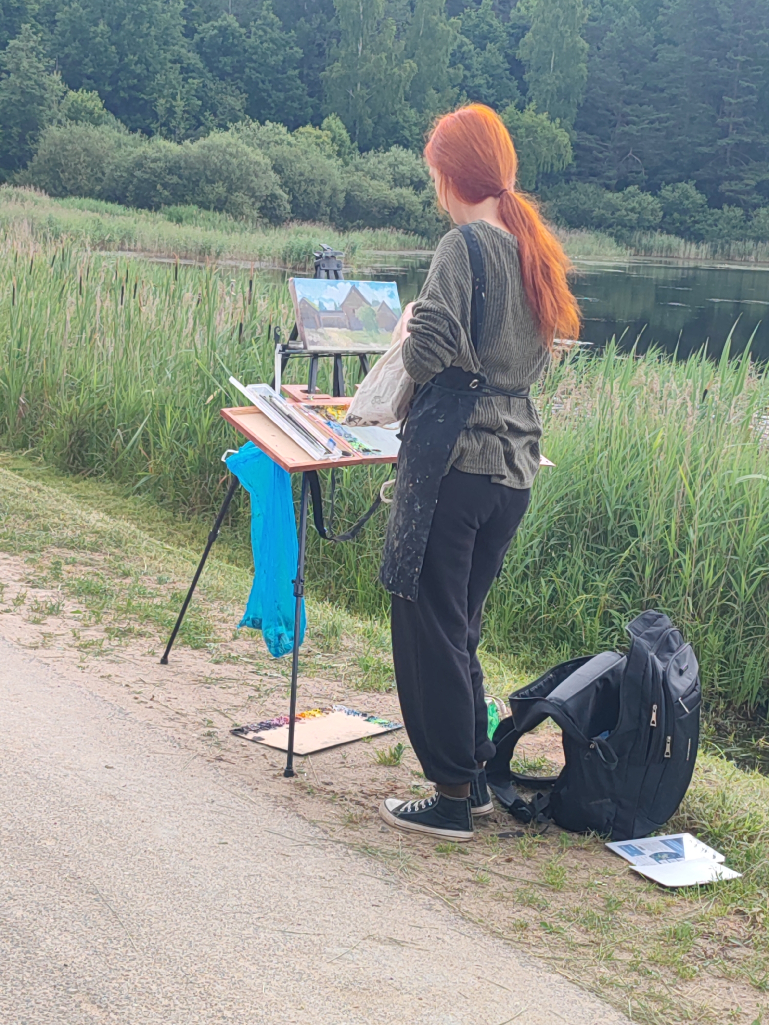 A woman with long, bright red hair tied in a ponytail stands facing away from the camera, painting a landscape on an easel. She is wearing a green, long-sleeved sweater, black pants, and black sneakers. A black apron is tied around her waist. Her art supplies, including a palette of colorful paints, brushes, and a blue plastic bag, are arranged on the easel and on the ground. A black backpack rests on the ground next to her, and some papers are spread out nearby. The scene is set in a natural environment with tall green grasses and reeds bordering a body of water. In the background, a dense line of trees forms a treeline against a hazy sky. The location is identified as Bugrovo, Russia.