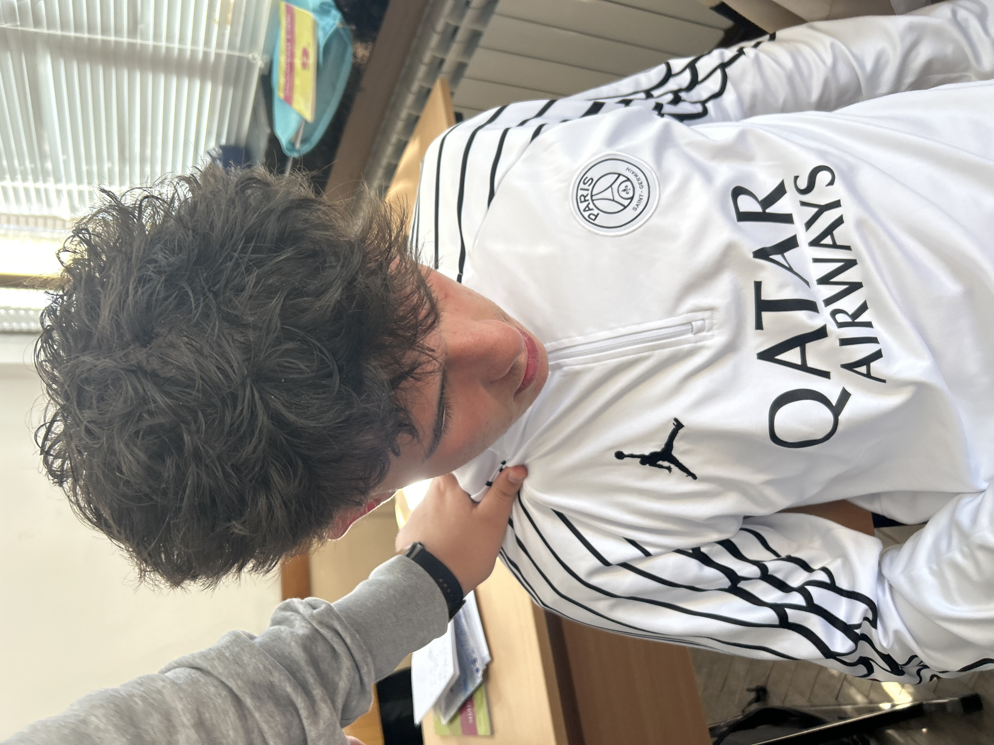 The image captures a close-up, angled view of a young person indoors in Zlatograd, Bulgaria. The main subject, likely male, has dark, curly hair and is shown from the chest up, looking downwards or to the side. They are wearing a white, long-sleeved athletic jacket with distinctive black stripes running along the shoulders and arms.
The jacket features prominent branding: a circular "PARIS SAINT-GERMAIN" club emblem is visible on the left chest area, accompanied by the text "QATAR AIRWAYS" written vertically in black capital letters. A black "Jumpman" (Jordan brand) logo is also present on the jacket sleeve.
Another person's arm and hand are visible in the foreground, gently resting on the main subject's shoulder. This hand is wearing a grey long-sleeved shirt and a black digital watch.
The setting appears to be an indoor space, possibly a room or classroom, illuminated by bright daylight suggesting it's daytime. In the background, hints of white vertical blinds or louvers are visible, along with a blue object and a yellow tag. A wooden surface, resembling a desk or table, is also partially seen with some papers or books on it. The main subject's face is partially obscured, making a clear emotional read difficult, but they appear calm. The interaction with the hand on the shoulder suggests a casual or friendly moment.