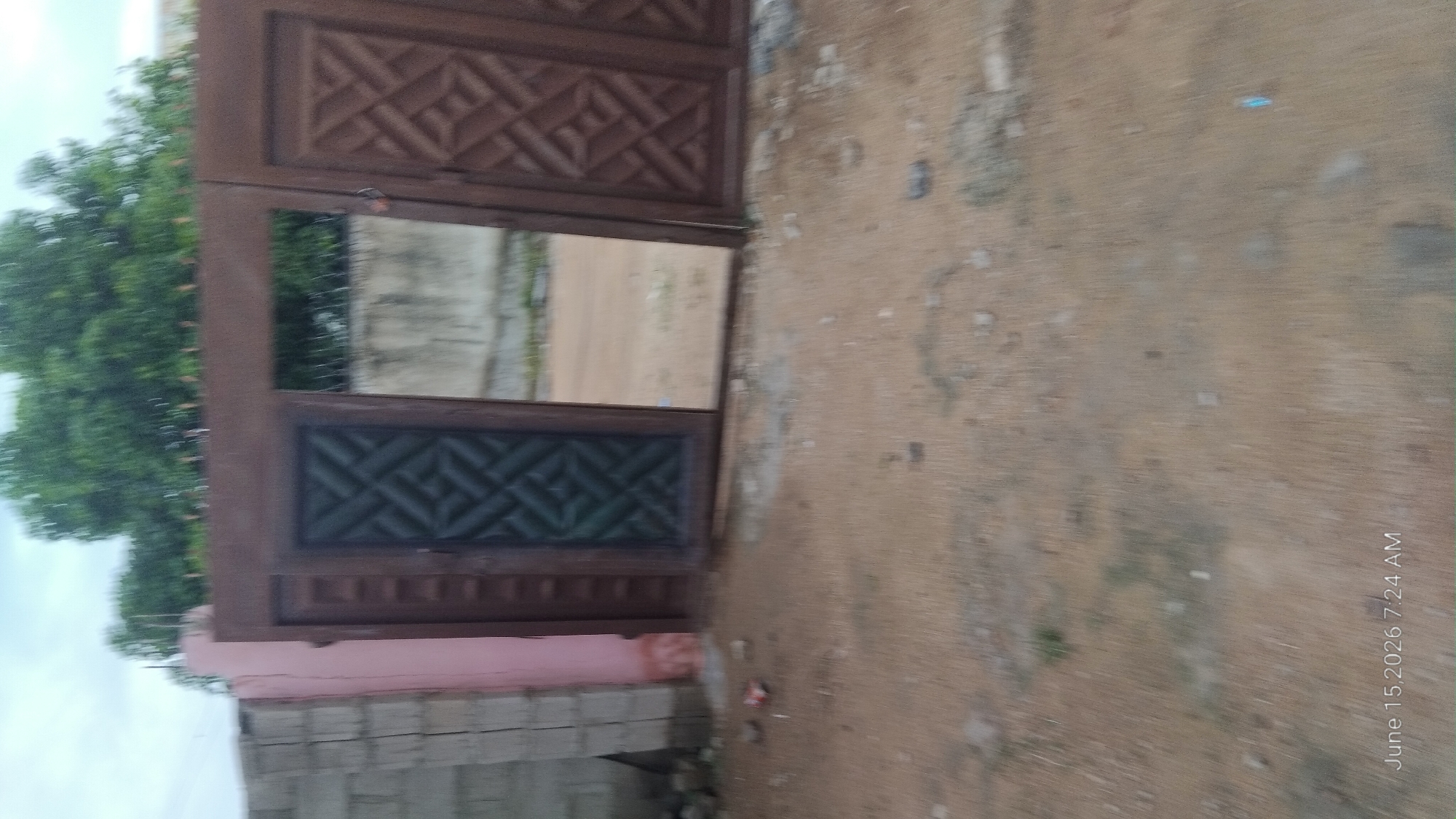 The image shows a brown metal gate with an open section, revealing a concrete wall and some greenery beyond. The gate has decorative patterns on its panels. To the left of the gate is a wall constructed of grey bricks. The ground in front of the gate is covered with dry, sandy soil. The scene appears to be outdoors, with a cloudy sky visible at the top of the image. The date and time stamp "June 15, 2026 7:24 AM" is visible in the lower left corner. There are no people visible in the image.