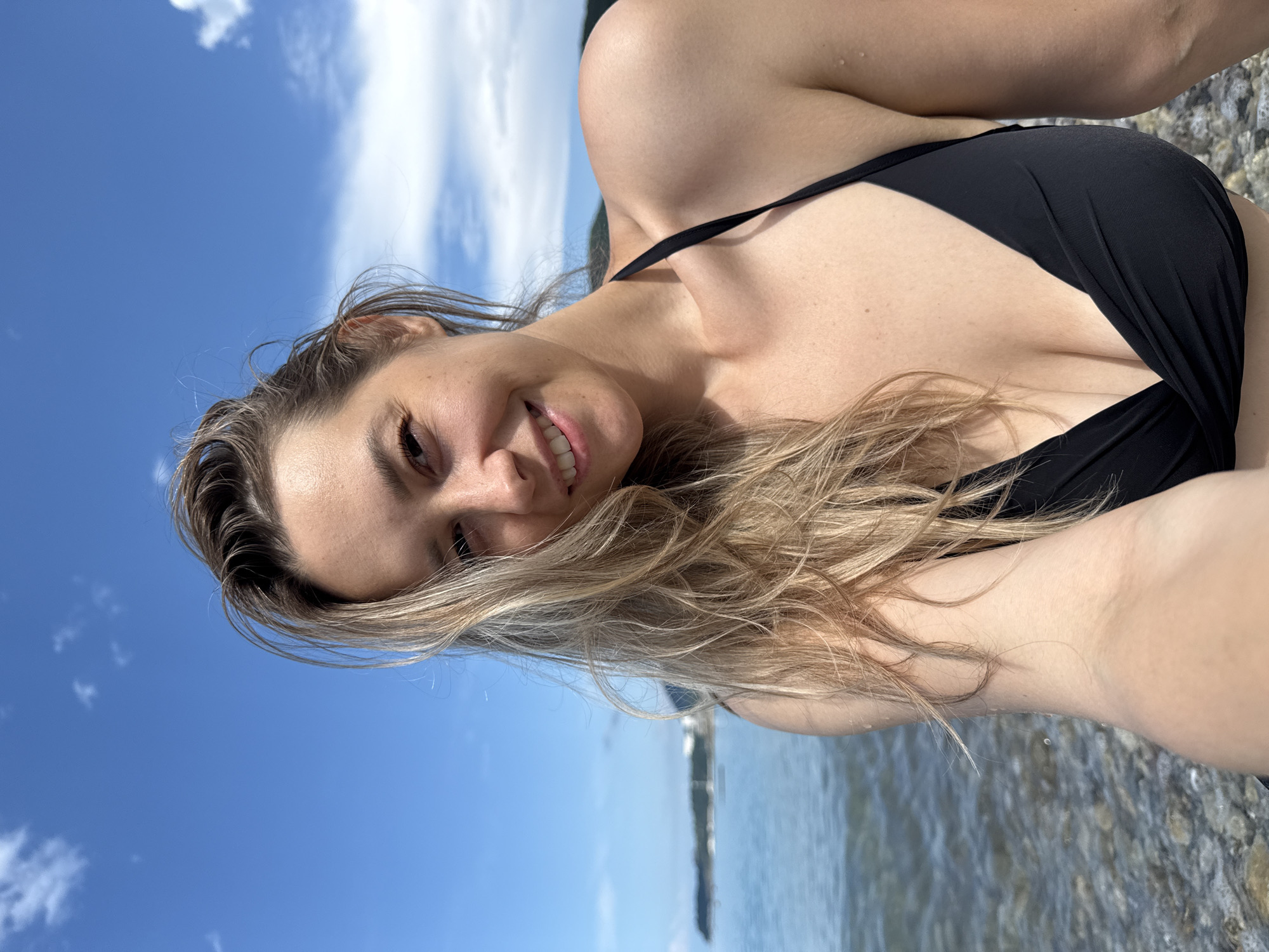 A woman with blonde hair is taking a selfie on a pebble beach. She is wearing a black bikini. The sea is calm and blue, with a shoreline and some greenery in the background. The sky is clear blue with a few white clouds. The location is the city of Nebug, Russia.