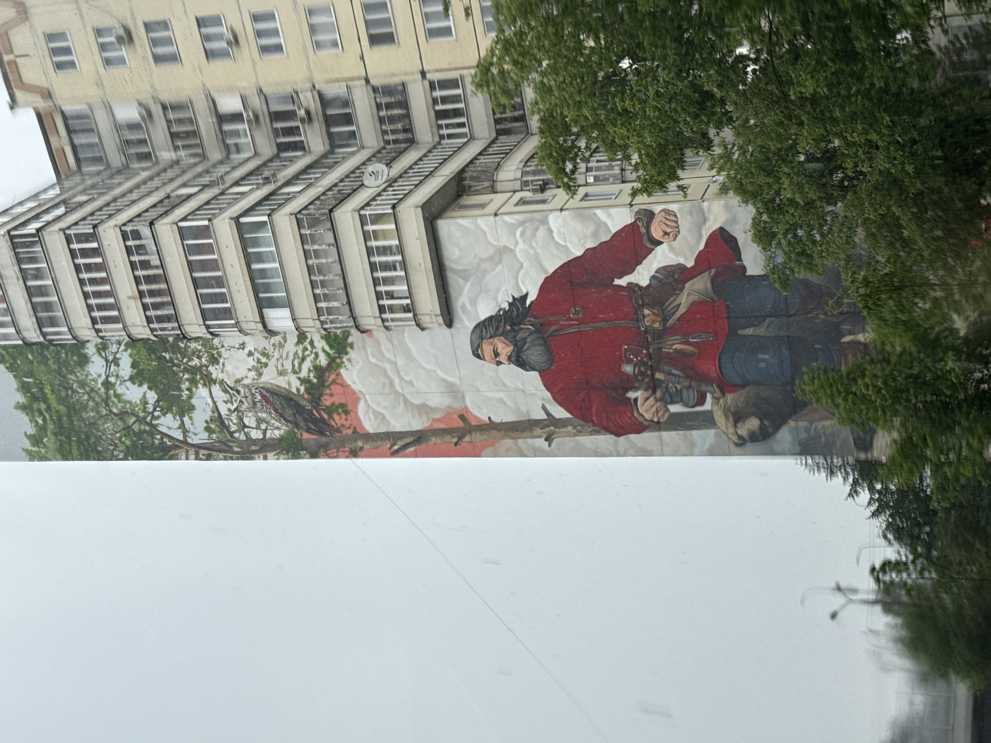 The image shows a mural painted on the side of a tall building in Perm, Russia. The mural depicts a man with a beard and long hair, wearing a red coat and blue pants, and holding what appears to be a camera. He is set against a backdrop of clouds and what could be a forest scene with a bird. The building has many windows and balconies. Trees partially obscure the mural on the right side of the image. The foreground shows an overcast sky.