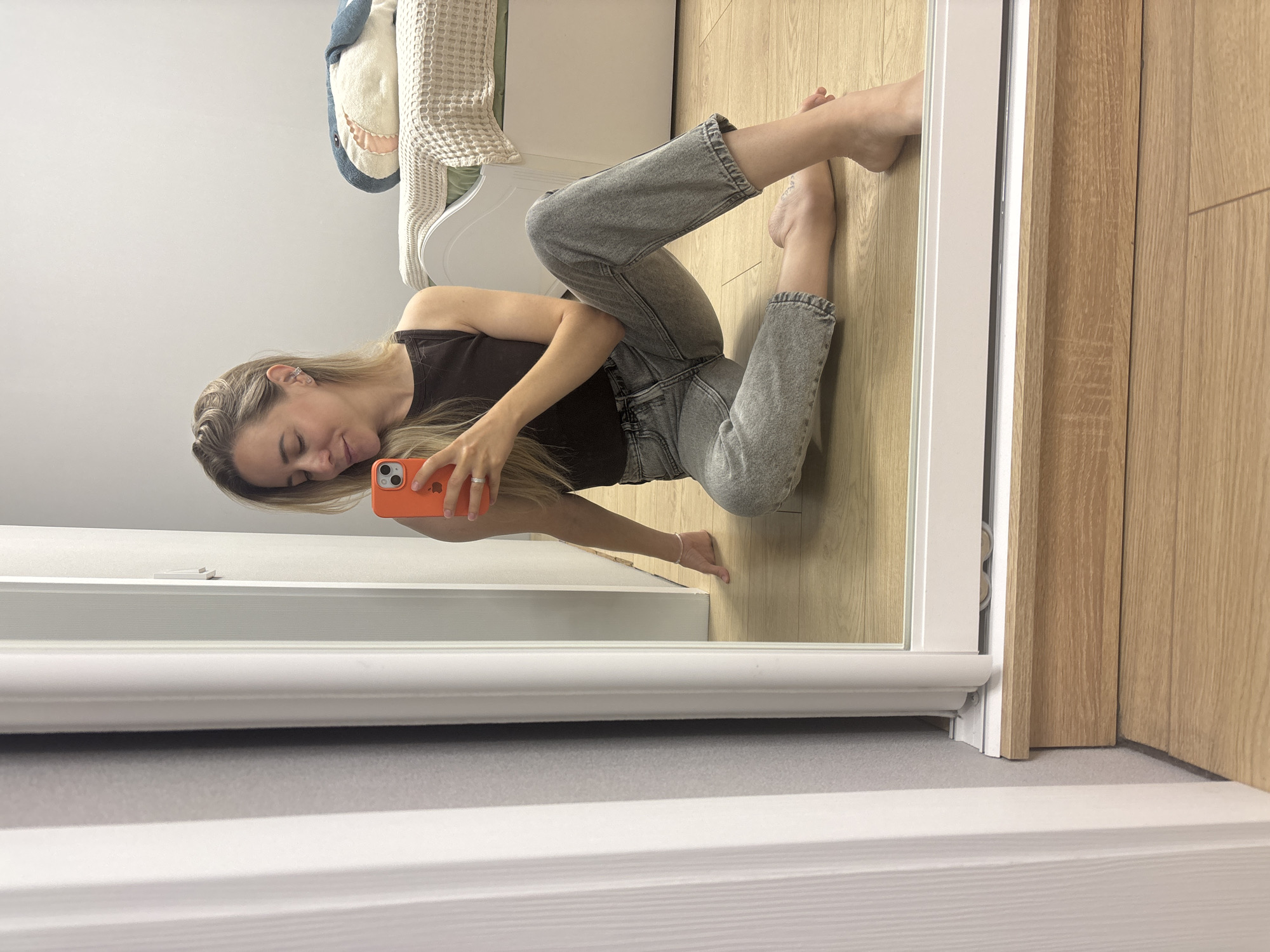 A blonde woman with fair skin, wearing a dark brown tank top and light gray jeans, is sitting on a wooden floor, taking a selfie in a mirror. She is wearing a silver ring on her left ring finger and a thin silver anklet. Her hair is partially pulled back, and she is wearing small stud earrings. A plush shark toy is visible on a bed behind her, which is covered with a cream-colored waffle-knit blanket and a light green sheet. The room appears to be a bedroom with light gray walls and white trim. The floor is made of light brown wooden planks. The scene suggests an indoor, domestic setting, likely in a private residence. The specific location in Chaikovsky, Russia, cannot be determined from the visual information provided.