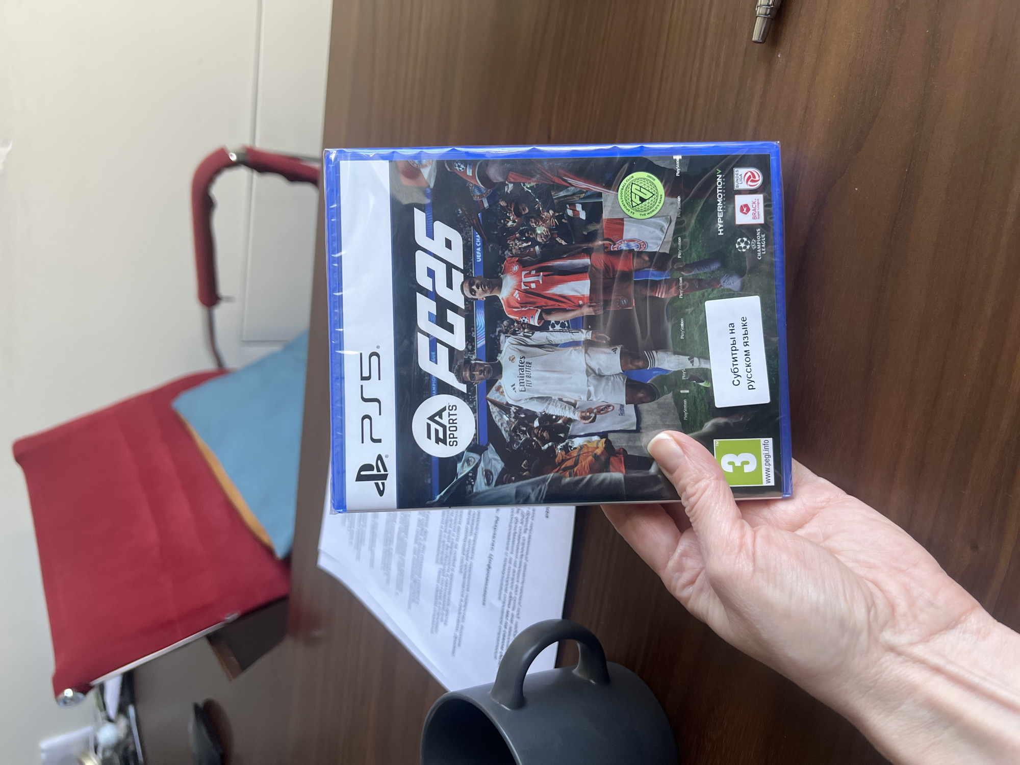 A person is holding a video game in their hand. The game is "FC26" for PlayStation 5. The box has a blue frame. The game is titled "Субтитры на русском языке", meaning "Subtitles in Russian". The PEGI rating is 3, indicating it is suitable for young children.
The game has the "HyperMotion V" title on the front.
The game features soccer players.
The game also has the EA Sports logo displayed on the front.
The background is a wooden table. In the background, there is a red chair and a blue cloth, plus some white paper. There is a gray mug near the bottom of the image.
The location is not discernible.