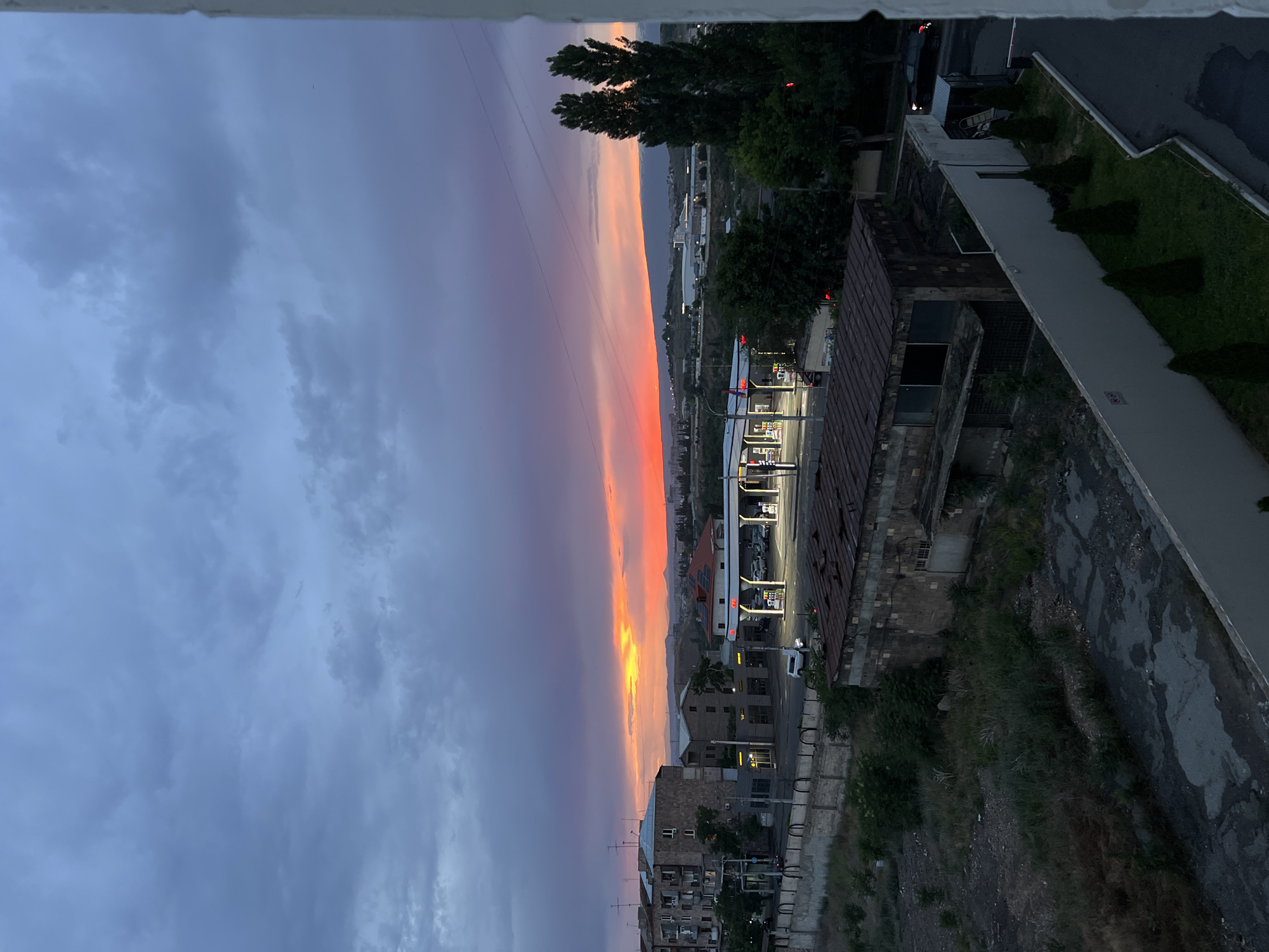 This detailed image captures an urban landscape in Yerevan, Armenia, during twilight, characterized by a vibrant sunset under an overcast sky.
The upper half of the frame is dominated by a cloud-filled sky. Heavy, dark blue-grey clouds occupy the zenith and upper portions, transitioning dramatically towards the horizon into a fiery band of bright yellow, intense orange, and deep red, which then fades into magenta and dark purple. Power lines or utility wires are visible against the sky in the mid-right section.
In the midground, a mixed-use environment is present. To the left, several light-brown, multi-story residential buildings, consistent with Soviet-era architecture, feature numerous television antennas on their rooftops. Below these buildings, a well-lit gas station with a large canopy illuminates several fuel pumps and parked vehicles, including a white SUV. Adjacent to the gas station, a small commercial structure is visible.
Below the street level, in the foreground-left, lies an undeveloped or abandoned plot of land. This area is overgrown with dry grass and sparse vegetation, interspersed with patches of dirt and gravel. Within this plot stands a dilapidated single-story building constructed from rough-hewn stone or brick, featuring a rusty corrugated metal roof and dark, empty window openings. A concrete retaining wall topped with dark metal railings separates the street from this lower-lying land.
To the right of the frame, a tall, white exterior wall runs diagonally, bordered by a strip of manicured lawn containing several conical evergreen shrubs. Beyond this wall, a dark paved area or service road is visible, with a dark car partially visible at the far right edge, displaying illuminated taillights. Two tall, slender, dark evergreen trees stand prominently in the mid-right, partially obscuring the distant view.
The background reveals an expanse of undulating hills or mountains beneath the colorful horizon, suggesting the city's geographical context. Scattered across these distant hills are various faint structures, appearing to be part of a further urban or industrial development. No clearly identifiable individuals are prominently featured, though faint figures may be present near the gas station pumps. The scene captures an intersection of established urban infrastructure, transitional land, and natural topography within the city of Yerevan.
