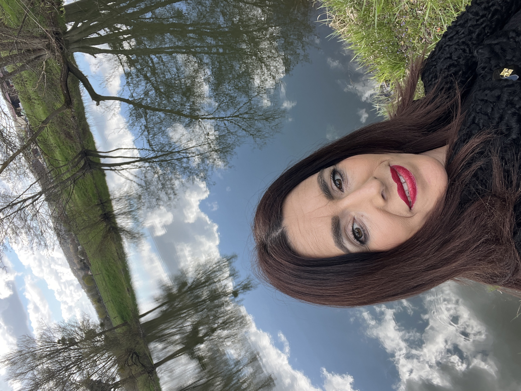 The image shows a woman with long, dark hair, wearing a black textured coat, posing for a selfie. She has fair skin, red lipstick, and appears to be smiling slightly. The background features a body of water reflecting the sky, clouds, and trees, suggesting the picture was taken near a river or lake. Based on the location provided, the scene is likely in Gavisse, France, where there are numerous waterways and natural landscapes. The environmental elements suggest an outdoor setting with calm waters mirroring the sky and surrounding vegetation.