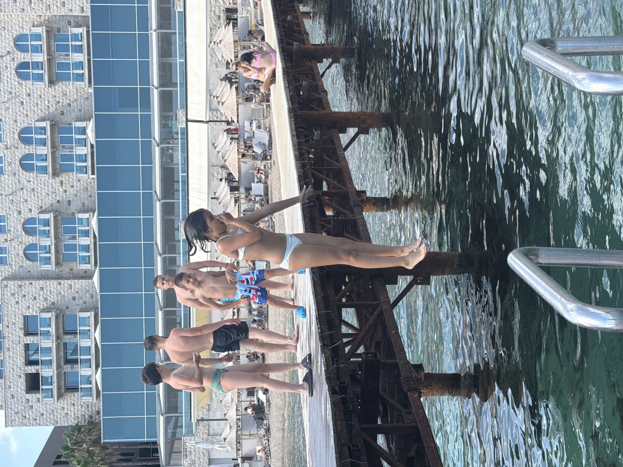 In this media file, a group of people are gathered on a wooden pier by the water. The pier is made of weathered wood and has rusted metal supports. There is a chrome railing on the right side of the frame. The water is clear and greenish, with ripples on its surface reflecting the sunlight.
In the foreground, a young woman in a white bikini is posing with her hand near her face. Behind her, a group of young men are standing on the pier, some in swim trunks. The pier is adjacent to a beach area with numerous straw umbrellas providing shade. In the background, there is a modern building with a facade of stone and blue glass windows.
The scene appears to be set on a sunny day, likely during the summer, given the attire of the people and the beach setting. The atmosphere seems relaxed and casual, with people enjoying the seaside. The location is identified as Budva, Montenegro. No visible text is present in the media file.