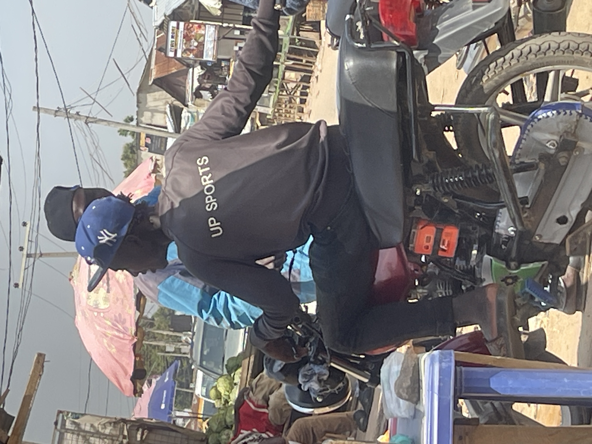 The image depicts a street scene in Abuja, Nigeria, with a person riding a motorcycle. The rider, wearing a black jacket with "UP SPORTS" printed on the back and a blue baseball cap, is the central focus. They are seated on a red motorcycle with visible parts like the engine, rear suspension, and wheel. To the left, a blue plastic table is present, and in the background, there are stalls with umbrellas and buildings, indicating a market or roadside commercial area. Other individuals are partially visible, suggesting a busy street environment. The overall setting appears to be outdoors during daylight, with a slightly overcast sky.