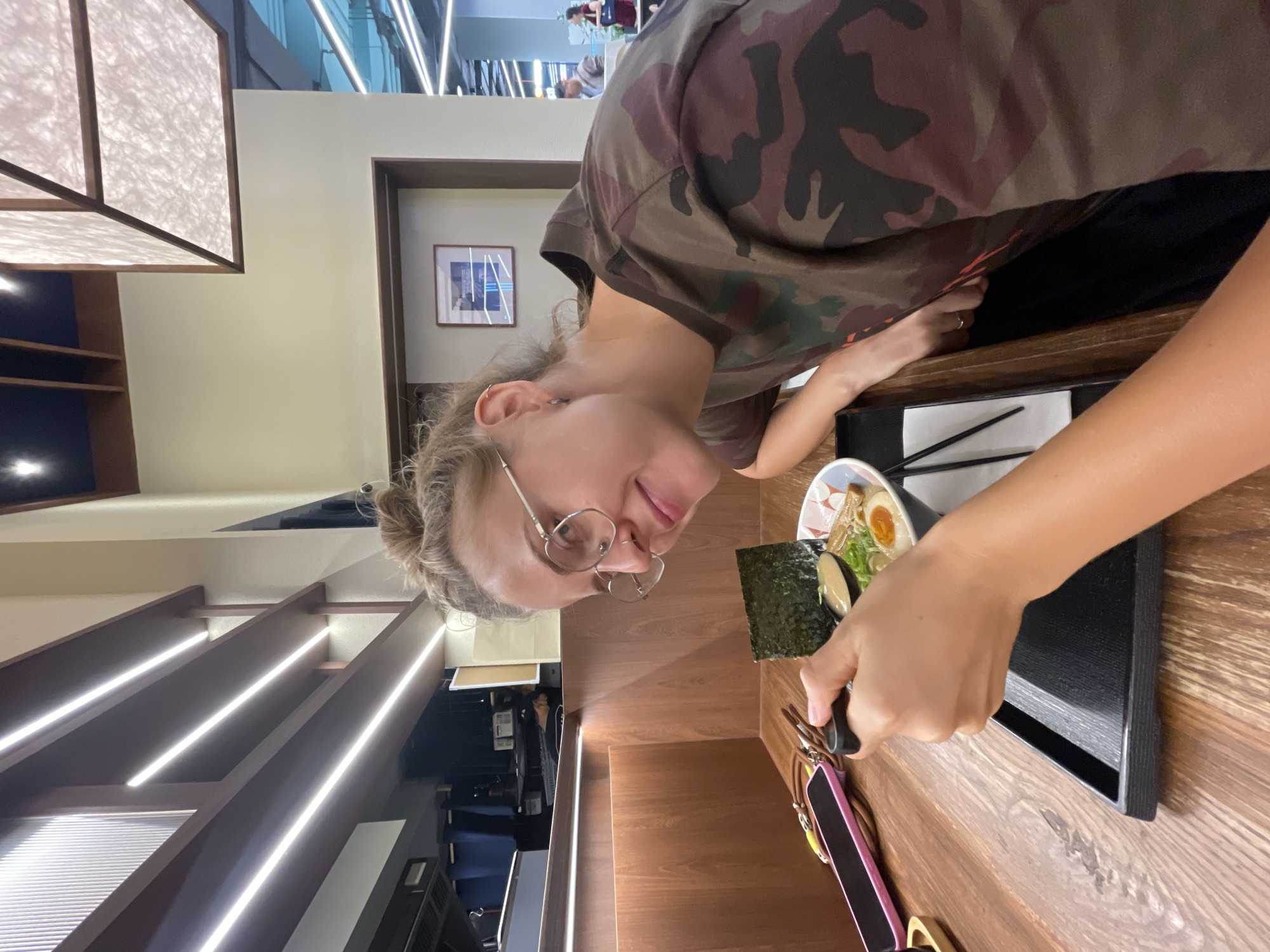 A woman with fair skin, wearing glasses and a camouflage T-shirt, is at a table in an indoor dining establishment. She is facing a bowl of ramen, which is set on a black tray. She is using a spoon and a sheet of seaweed to prepare a bite. The table is wooden. A pink and brown item is on the table to her left. The background shows architectural details, including lighting fixtures and part of another dining level. The location is likely in Bangkok, Thailand.