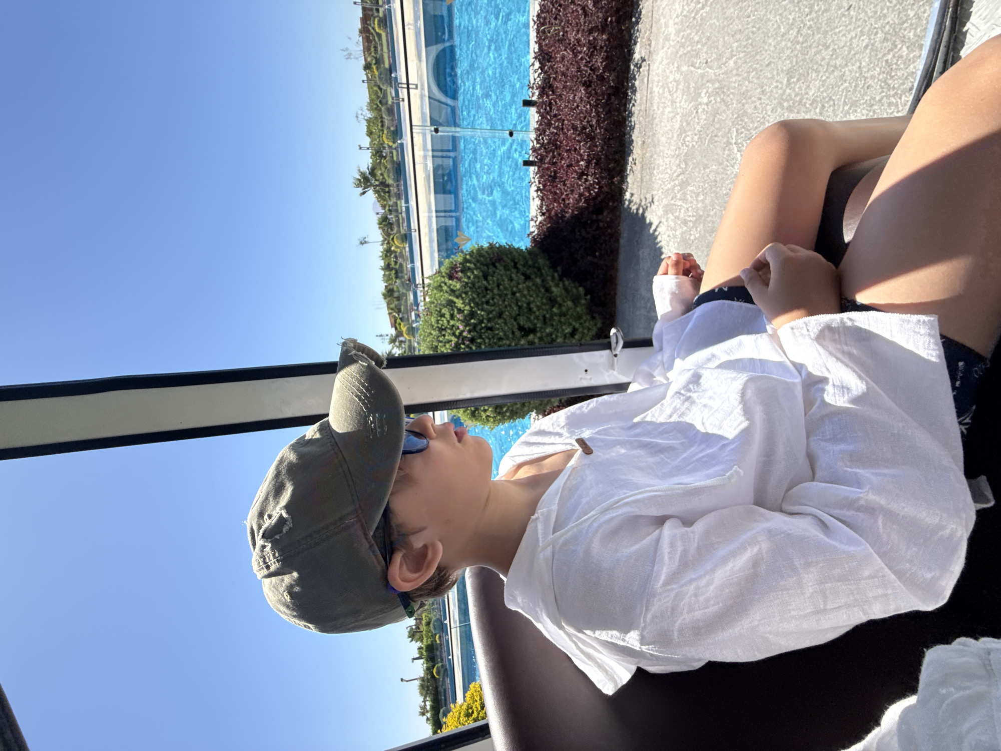 A boy wearing a green baseball cap and sunglasses is seated in a vehicle. He is wearing a loose white hooded shirt. The vehicle appears to be moving, as indicated by the blurred background. Outside the vehicle, a bright blue swimming pool is visible, surrounded by lush green bushes and red-leaved plants. Palm trees and other vegetation are seen in the distance, under a clear blue sky. The setting suggests a sunny day in a resort or park. Given the context of Sharm El Sheikh, Egypt, this could be a scene at a hotel, a water park, or a similar recreational facility.
