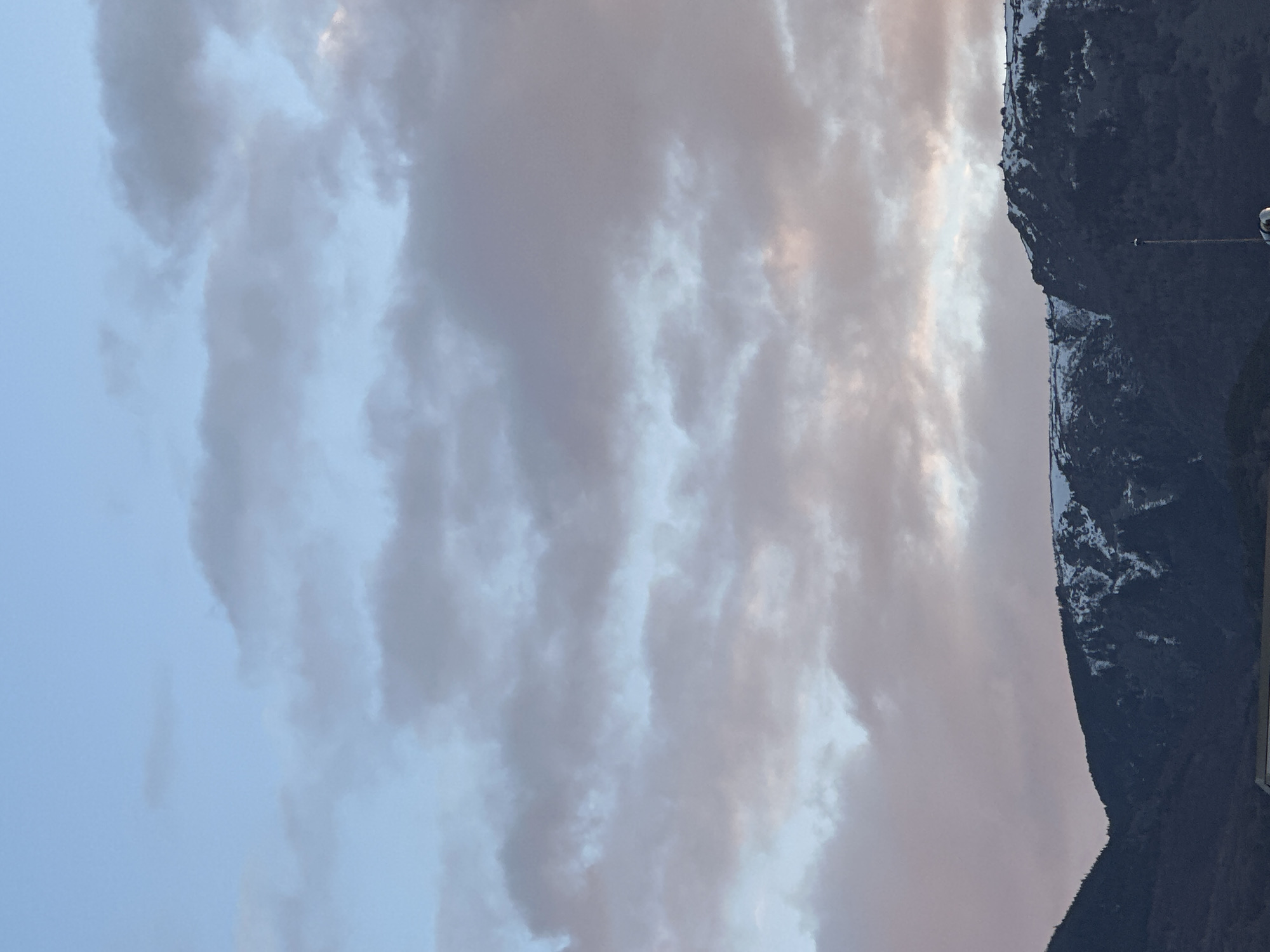 The image captures a serene mountainous landscape under a cloudy sky, likely at either sunrise or sunset. The scene is dominated by a vast expanse of sky in the upper two-thirds of the frame and a dark mountain range forming the lower third.
The sky is largely covered by layered clouds, displaying a blend of soft blue, grey, pink, and pale orange hues. These colors suggest diffused light, typical of twilight hours. The clouds are dense but allow some glimpses of lighter, clearer sky, particularly towards the left.
The mountain range, possibly part of the Vitosha mountains near Sofia, Bulgaria, appears as a dark, rugged silhouette against the lighter sky. Its peaks are partially covered with patches of snow, indicating a colder season. The mountain slopes are steep and appear forested, though details are obscured by the low light and distance. Along the ridge line, a slender, dark structure resembling a utility pole or antenna mast is faintly visible on the right side.
No people or explicit activity are present, emphasizing the quiet majesty of nature. The overall impression is one of peacefulness and natural grandeur, captured during a transient moment of natural light. There is no visible text in the image.
