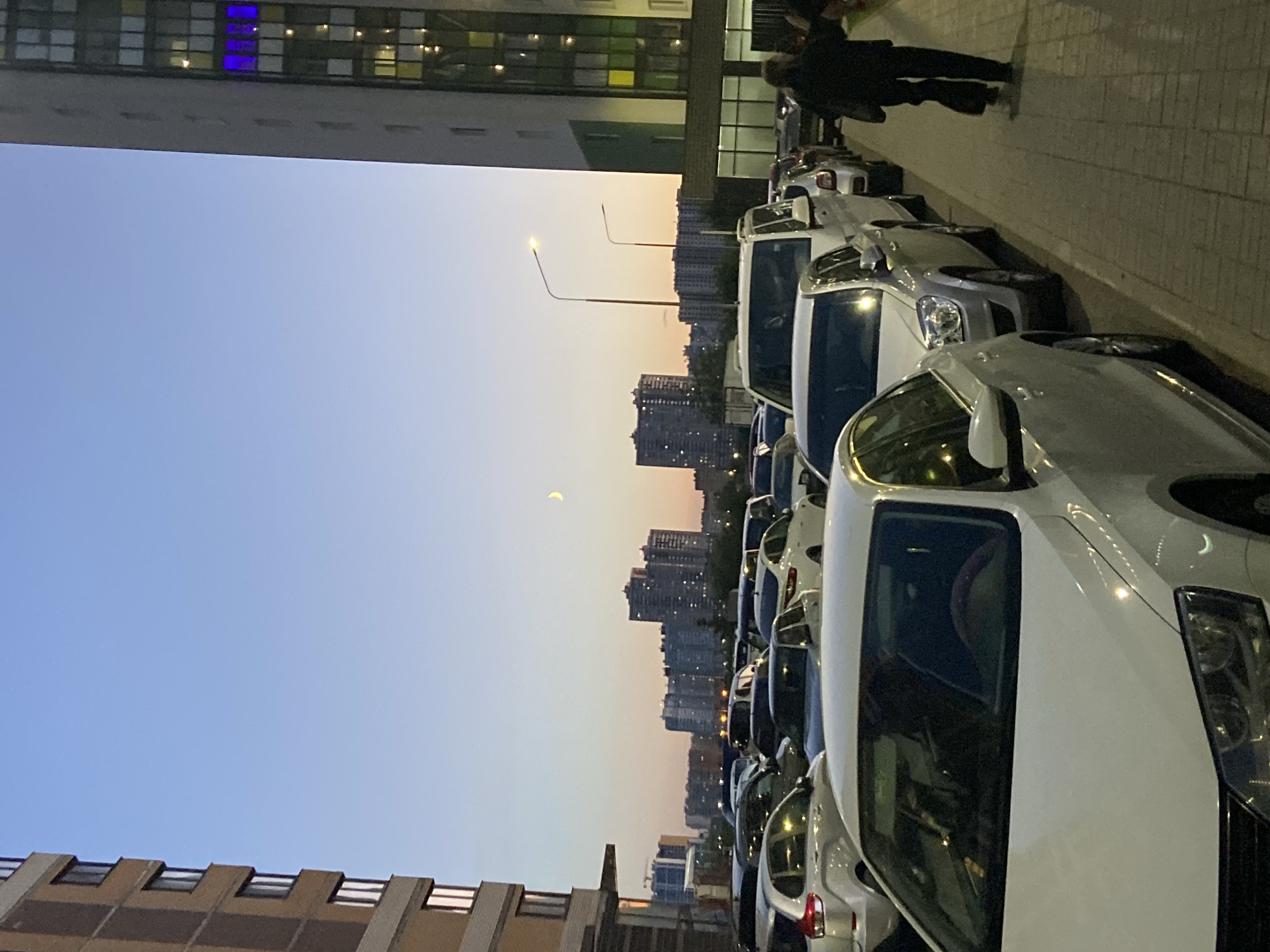 A low-angle, outdoor, eye-level shot captures a residential parking lot at dusk in an urban environment, likely in Russia, given the context. The sky is a gradient of pale blue transitioning to soft orange and pink near the horizon. A sliver of a crescent moon is visible in the upper center of the sky.
Dominating the left side of the frame is the facade of a modern, multi-story apartment building, constructed with light brown brick and off-white concrete sections, featuring numerous windows. To the right, another taller apartment building with a predominantly white facade and dark windows is visible. One section of this building emits a vibrant blue light from a cluster of windows.
The parking lot is densely packed with a variety of cars, mostly sedans and SUVs, parked in rows. The cars are predominantly white and silver, with some darker vehicles interspersed. The lighting suggests it is nearing nighttime, with streetlights illuminating the scene and lights on within the apartment buildings.
In the lower right quadrant of the image, a person is walking away from the camera along a paved sidewalk that runs alongside the parking lot. The person is wearing dark clothing, including a jacket and pants, and carrying a dark bag over their shoulder. The sidewalk is made of light-colored paving stones.
In the background, more apartment buildings of varying heights are visible, forming a dense urban skyline. Some of these buildings also show illuminated windows. Two streetlights with single lamps on tall poles are present in the mid-ground. The overall impression is a typical residential area in a city during the evening transition.