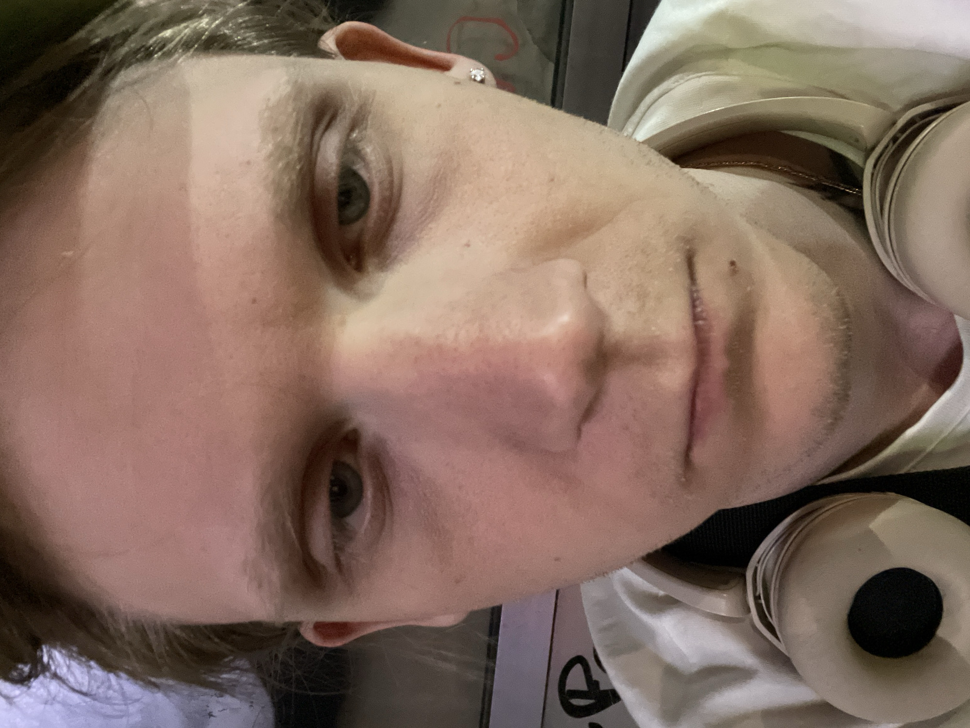 A close-up, high-angle shot of a fair-skinned individual with short blonde hair. The person has light green eyes, a prominent nose, and stubble. They are wearing a white t-shirt and have headphones around their neck. The location is undetermined. There is no reliable information in the image to specifically determine that the image was taken in the city of Palilula Urban Municipality, Serbia.