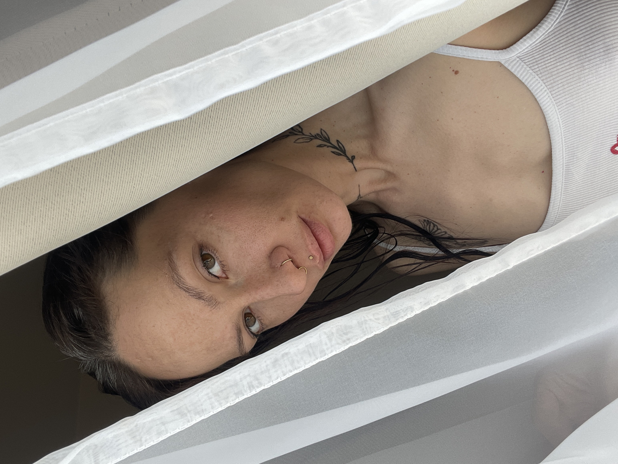 A woman is captured in an unusual, angled perspective. The photograph is framed by a layer of sheer white fabric in the foreground and a thicker, beige fabric behind her. She is looking directly at the camera with a slight tilt of her head. The visible portions of her body reveal she is wearing a white ribbed tank top. She has a tattoo of a leafy branch design on her neck. She is wearing multiple pieces of jewelry, including a ring in her nose.
There is nothing in the photo to indicate that it was taken in Izhevsk, Russia.