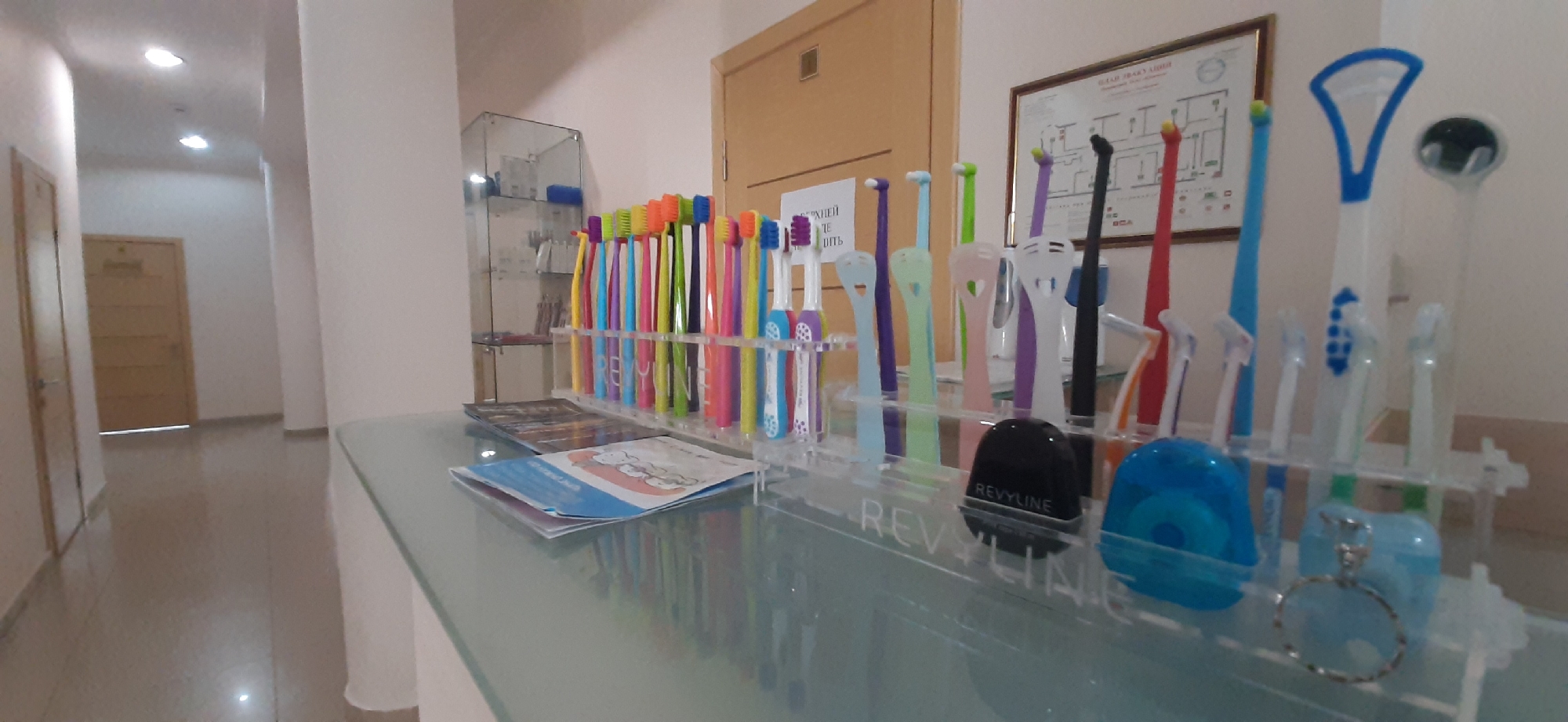 The image depicts a reception desk in what appears to be a dental clinic or similar healthcare facility in Yessentuki, Russia. The desk is made of glass and reflects the surrounding environment. Arranged on the desk are numerous colorful toothbrushes of various brands and styles, including some specialized tongue cleaners. A dispenser of dental floss, a small black circular object, and other oral hygiene products are also visible. In the background, a doorway with a wooden door is present, and a hallway extends further into the building, with lighting fixtures on the ceiling. A framed diagram, possibly a floor plan or emergency evacuation map, hangs on the wall above the reception desk. A glass display cabinet containing additional dental products is positioned to the left of the desk. The overall impression is one of a clean and organized professional setting focused on dental care.