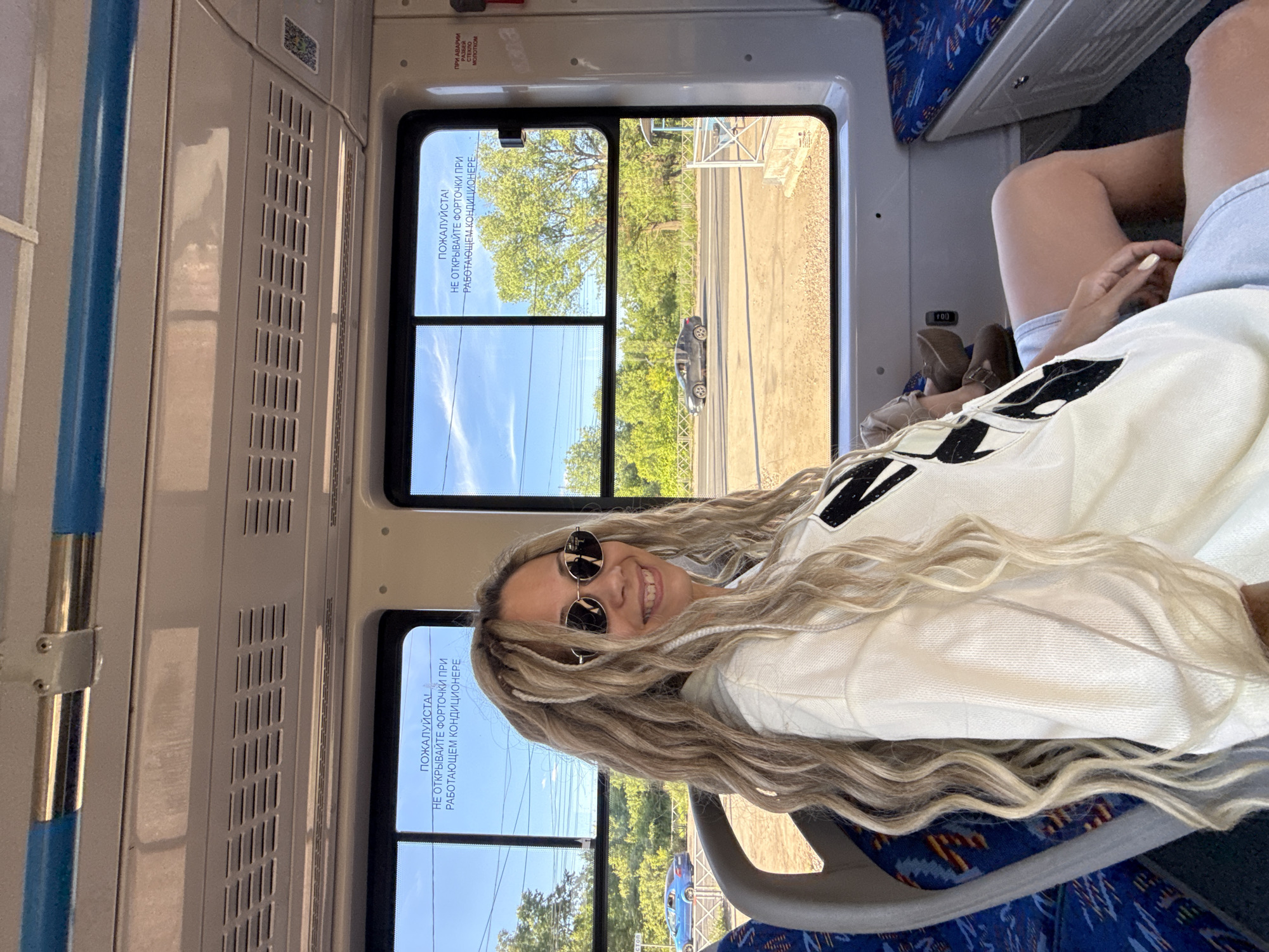 A woman is seated inside a public transport vehicle, possibly a train or electric multiple unit, on a sunny day in Rusanovo, Russia. The woman, with long, wavy blonde hair featuring some dreadlocks or braids, is wearing sunglasses, a white long-sleeved top with a black graphic design, and light-colored shorts. She is smiling and looking directly at the camera. Her fingernails are painted with a light polish.
The vehicle's interior features blue-patterned seats, a white ceiling with air vents, and overhead blue handrails. Through the large windows, an exterior scene is visible: a clear blue sky with some clouds, a paved road, and abundant green trees and foliage suggesting a suburban or rural environment. On the road, a blue sedan is visible on the left side of the frame, and a dark-colored sedan is further down the road on the right. A fence runs along the roadside, and a small shelter-like structure is partially visible on the far right.
Text is prominently displayed on the windows in Russian. It reads, "ПОЖАЛУЙСТА! НЕ ОТКРЫВАЙТЕ ФОРТОЧКИ ПРИ РАБОТАЮЩЕМ КОНДИЦИОНЕРЕ." which translates to "PLEASE! DO NOT OPEN THE VENTS WHEN THE AIR CONDITIONER IS WORKING." A partially visible emergency sign on the far right window states, "ПРИ АВАРИИ РАЗБЕЙ СТЕКЛО МОЛОТКОМ," meaning "IN CASE OF EMERGENCY BREAK GLASS WITH HAMMER."