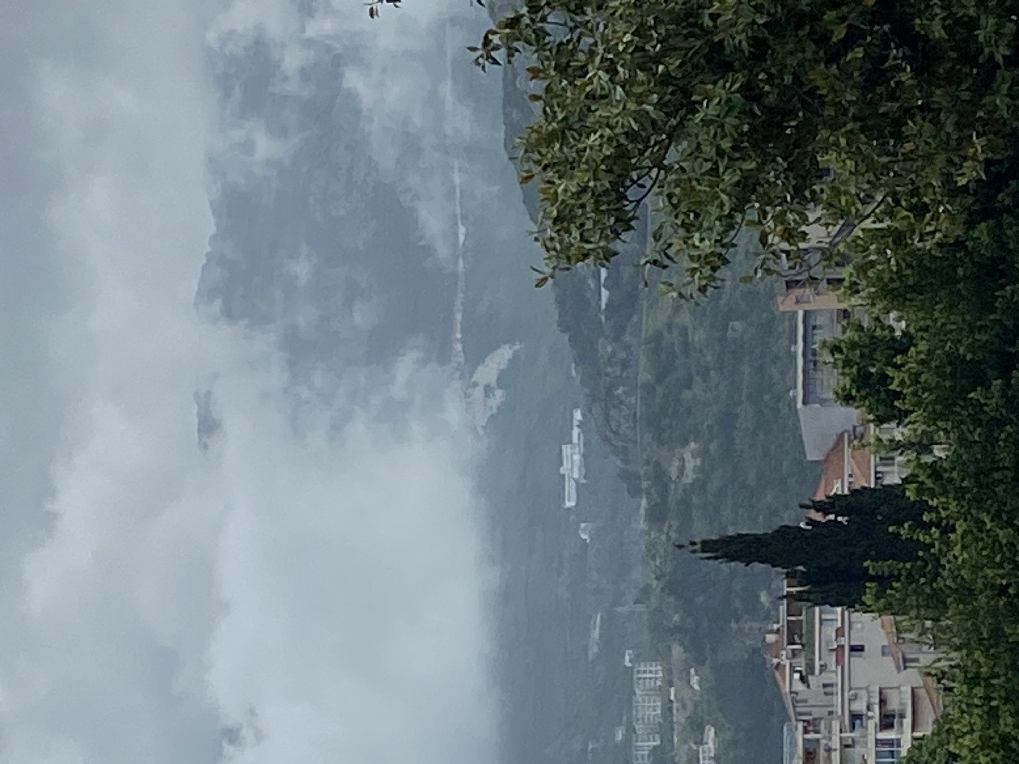 The image shows a view from Budva, Montenegro, featuring buildings and a mountain covered in fog. The foreground contains greenery, partially obscuring the buildings. In the background, a mountain is heavily obscured by fog. The scene suggests a cloudy or overcast day.
