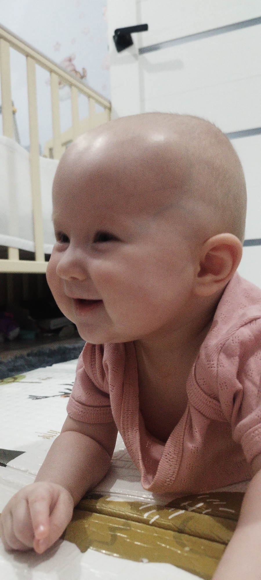 A baby is lying on a white mat with geometric patterns. The baby is wearing a pink knit shirt and is facing to the left with a slight smile. A crib is visible in the background, with a white wall and a black fixture above it. The location is not identifiable as Yakutsk, Russia based solely on the image.