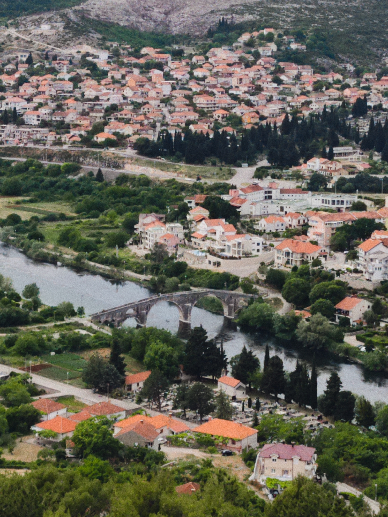 The image shows an aerial view of Trebinje, Bosnia and Herzegovina, featuring the Arslanagić Bridge. The Neretva River flows through the scene, with the stone arch bridge spanning it. Buildings with orange-tiled roofs and surrounding greenery characterize the urban landscape. The terrain slopes upwards towards a hill or mountain in the background. The bridge has multiple arches supported by pillars. A road follows the river's edge on both sides, connecting to the bridge. Buildings with mixed architectural styles and sizes are clustered along the river and extend towards the top of the image. Cypress trees are visible near the river and among buildings.