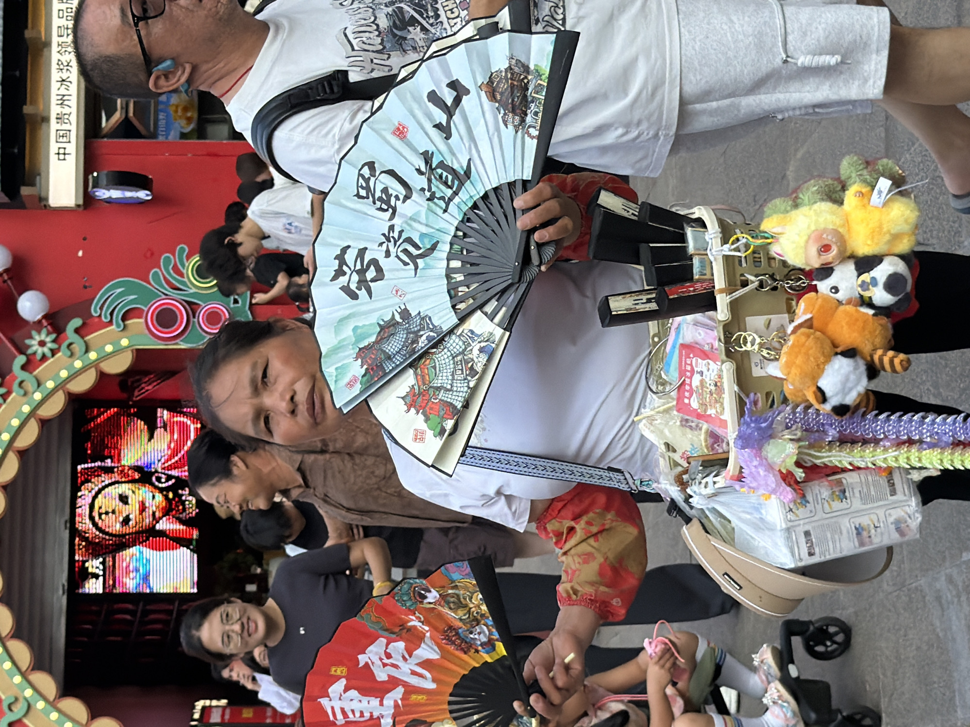 In Yuzhong District, China, a woman is holding two traditional folding fans, one red and one light blue, each adorned with Chinese calligraphy and painted scenes. She wears a white shirt and a red-patterned arm covering. A basket filled with various items, including stuffed animal keychains, is attached to her. In the background, a man in a white t-shirt and grey shorts is visible, as well as other people in a bustling urban environment with decorated archways and signs.