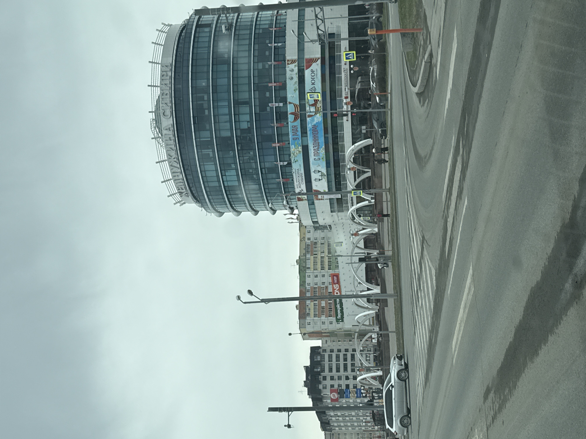 The image captures a street scene in Tobolsk, Russia, presenting an urban landscape on a cloudy day. Prominently featured is a modern, cylindrical building with a glass facade; its name, "Zhemchuzhina Sibiri," or "Pearl of Siberia," is written at the top. Banners hang along the building, including one mentioning "9 Maya" (May 9th) and another reading "S prazdnikom!" (With the holiday!). Below the modern building, older apartment buildings line the street. A crosswalk spans the road, and pedestrian bridges or artistic installations are visible alongside the buildings. Several cars are parked on the side of the road. Streetlights are present, and the sky is overcast.