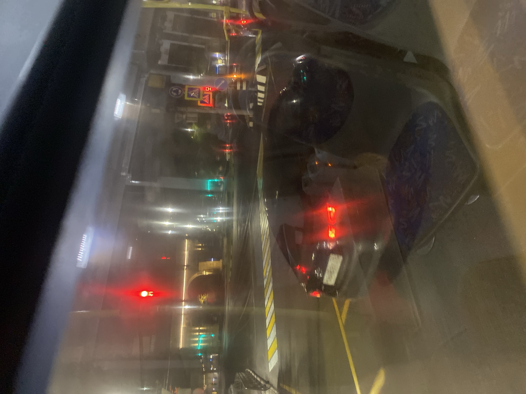 The image, possibly taken from inside a vehicle, shows a street scene at night in what appears to be Заволжье, Russia. Streetlights illuminate the road, which has yellow and white painted lane markings. Traffic lights are visible with red and green signals. Cars are on the road, and a vehicle with red tail lights is in the foreground. Street signs are present along the roadside. Through a wet or dirty windshield, the streetscape is somewhat blurred.