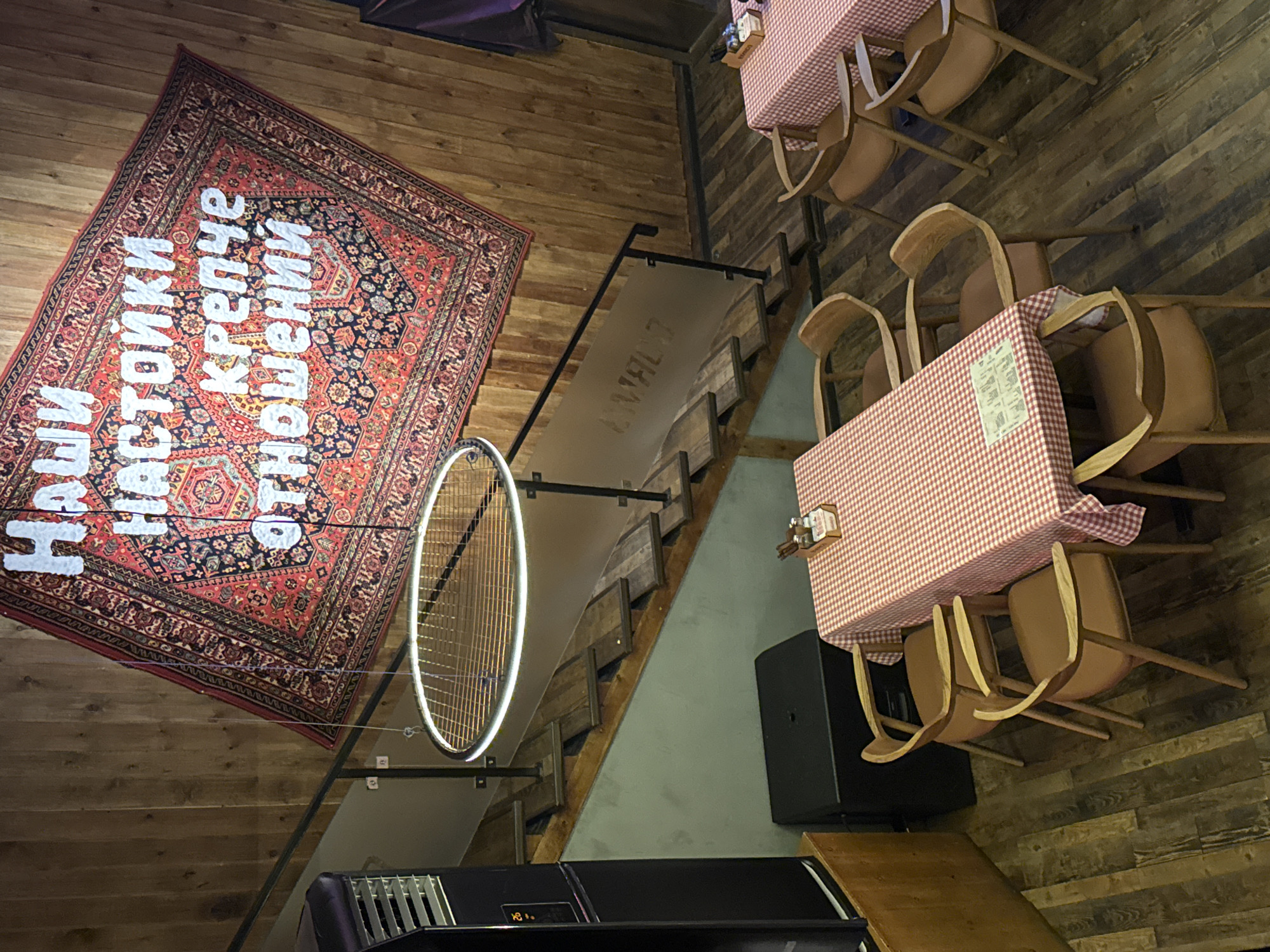 The image shows a multi-level restaurant interior, possibly in Tashkent, Uzbekistan, judging by the Cyrillic text on a large carpet hanging on a wall. The restaurant features wooden flooring and wooden stairs leading to different levels. Two tables are visible, each covered with a red and white checkered tablecloth and surrounded by wooden chairs. A circular, illuminated light fixture hangs near the stairs. A black speaker is situated near one of the tables. The overall lighting is dim, creating a warm ambiance. The architectural style appears modern with rustic touches.