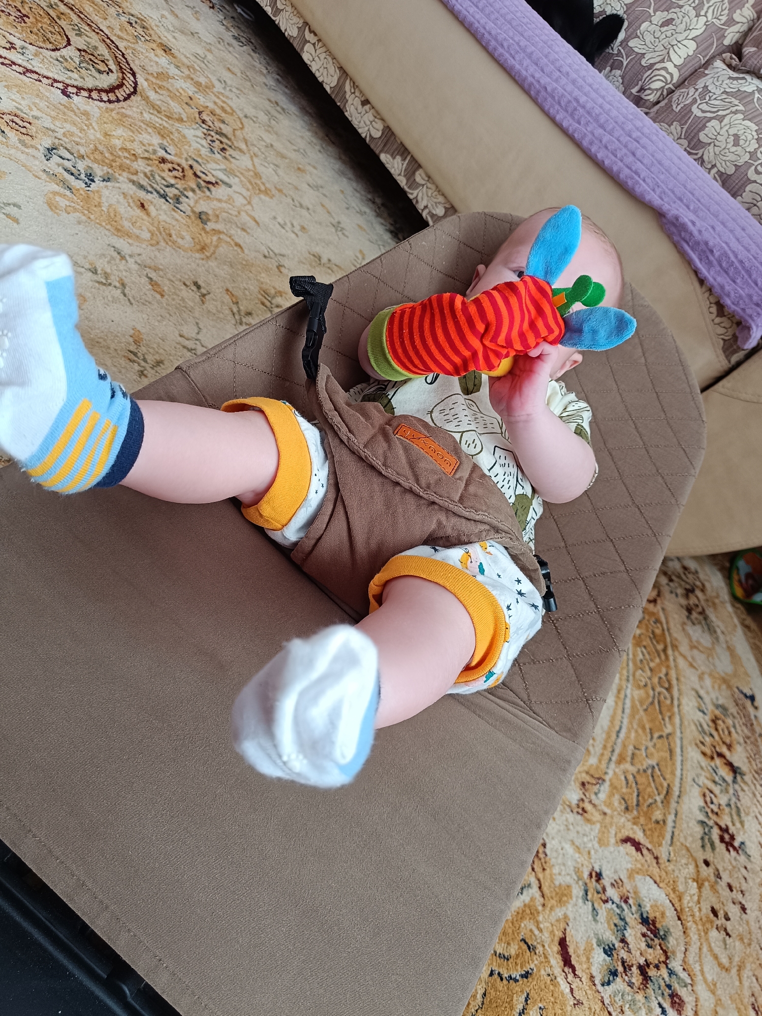 A baby lies in a brown baby carrier with quilted stitching. The baby wears a patterned white and green shirt, brown shorts with orange trim, and mismatched socks (one white and blue with a yellow stripe, the other white with a blue toe). The baby is holding a red, orange, and blue stuffed toy. The carrier is placed on a patterned beige and brown carpet. Part of a beige sofa is visible behind the baby. The location is Tobolsk, Russia.