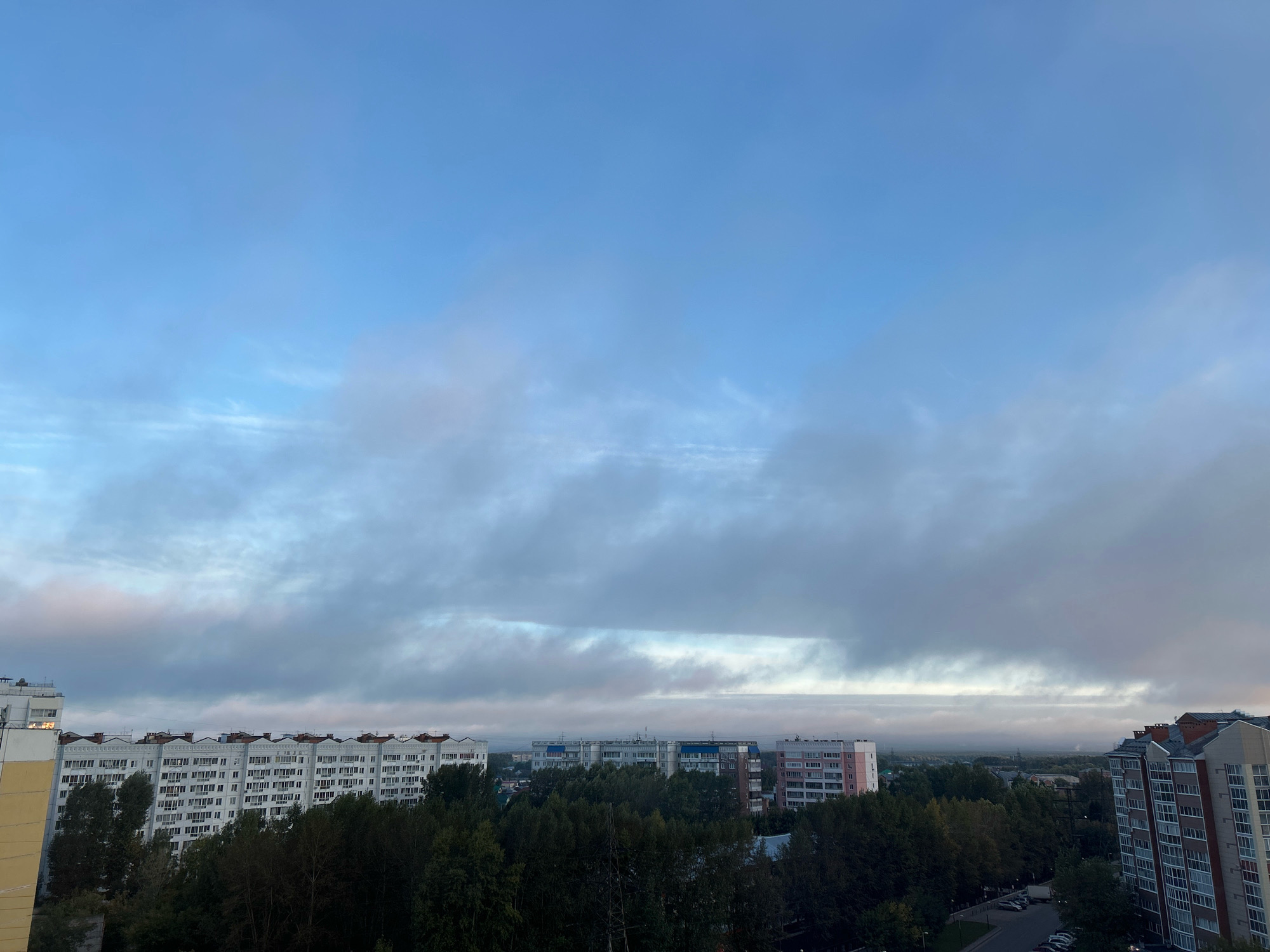 The image depicts a broad view of a city, likely Tomsk, Russia, under a cloudy sky. The sky is a pale blue with scattered, elongated clouds, suggesting either early morning or late afternoon. Below the sky, a dense line of trees forms a verdant canopy over a residential area. Several multi-story apartment buildings are visible above the trees. On the left, a white and yellow building stands prominent. In the center, a long, white apartment building with a rhythmic pattern of windows and balconies stretches across the frame. Further to the right, a pink and white building and a darker, more modern building with balconies are visible. A street with parked cars can be seen to the right of the buildings. The overall impression is one of a typical urban landscape in a temperate climate. There are no people or animals visible in the image.