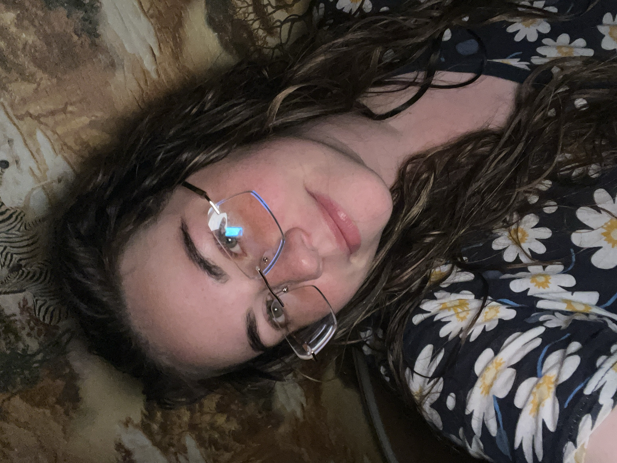 This image depicts a female subject in an indoor setting, identified as being in Сосново, Russia. The close-up selfie captures a woman with long, wavy, dark brown hair featuring lighter highlights, flowing over her shoulders. She has a fair complexion, hazel-colored eyes, and dark, well-defined eyebrows. Her lips are full, slightly parted in a gentle smile, and appear glossy. She is wearing rimless or semi-rimless rectangular glasses; the left lens (viewer's right) exhibits a distinct blue/cyan reflection, suggesting illumination from a screen. Her attire is a dark-colored top patterned with white and yellow daisies, including subtle blue accents. The background behind her is a textured surface, likely fabric or wallpaper, featuring an African savanna theme with light brown and beige tones, dark brown tree silhouettes (resembling acacia trees), and prominent black and white zebra stripes visible in the upper left. The scene is dimly lit, with the subject's face being the most illuminated area, likely by the light source causing the reflections in her glasses.