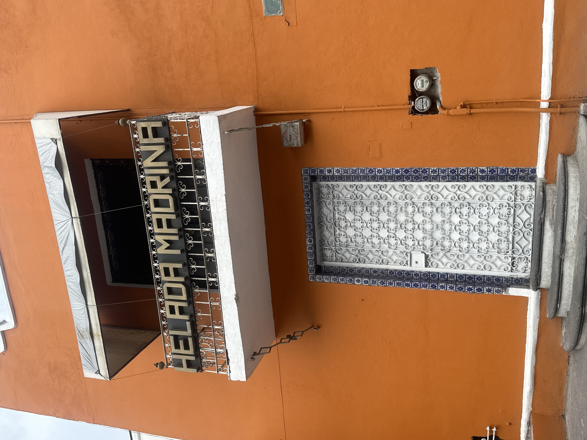 The image shows the exterior of a building with an orange facade in the city of Unknown, Unknown. The building features a metal sign reading "HELADA MADRINA" mounted above an opening. A decorative, white, wrought-iron gate, framed with blue and white tiles, is situated next to a stairway. An electrical box is visible on the upper portion of the wall, with exposed meters.