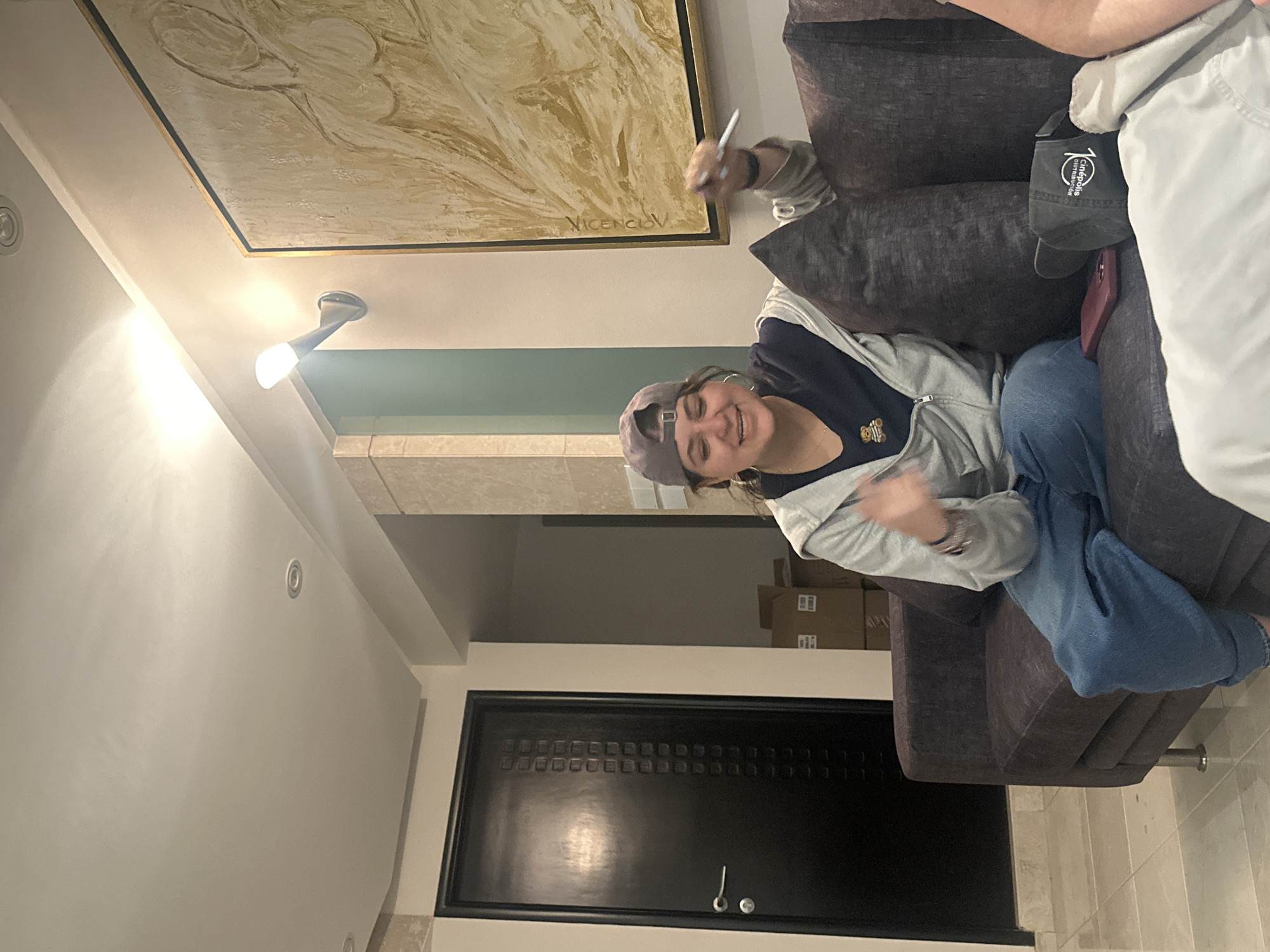 A person wearing a gray baseball cap, gray hoodie, and blue jeans is sitting on a gray couch in an interior setting. This person is holding a pen or marker in their right hand and appears to be signing or writing on an artwork hanging on the wall. The artwork is a large, abstract painting in shades of beige and brown with a gold frame. A second person is partially visible to the right, wearing a light-colored shirt and a dark-colored baseball cap. The background includes a hallway, a black door, and boxes stacked in the distance. There is a sconce visible on the wall. The location is Mexico City, Mexico.