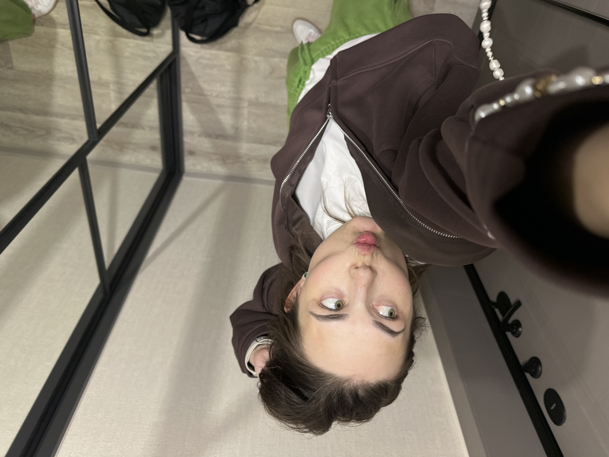 A young female, likely in Perm, Russia, takes a playful selfie indoors. She is positioned centrally, looking upwards with her head tilted and making a "fish face" expression with puckered lips. Her wide, green-brown eyes look slightly to the side. She has dark brown hair and is wearing a dark brown hooded zip-up jacket over a white top. A pearl-like beaded strap or necklace is visible around her neck/shoulder, and a small green earring adorns her left ear. A silver-toned watch or bracelet is on her left wrist.
In the background, light-colored wooden or laminate flooring is visible, along with black metal frames, possibly part of a room divider, shelving unit, or mirror structure. To the right, a light-colored wall or door features black door handles and a lock. Faintly visible in the upper left background are two black bags. The setting appears to be a modern indoor space. The lighting is bright and even, consistent with daytime or well-lit artificial interior lighting. The image is rotated 90 degrees clockwise. No visible text is present.