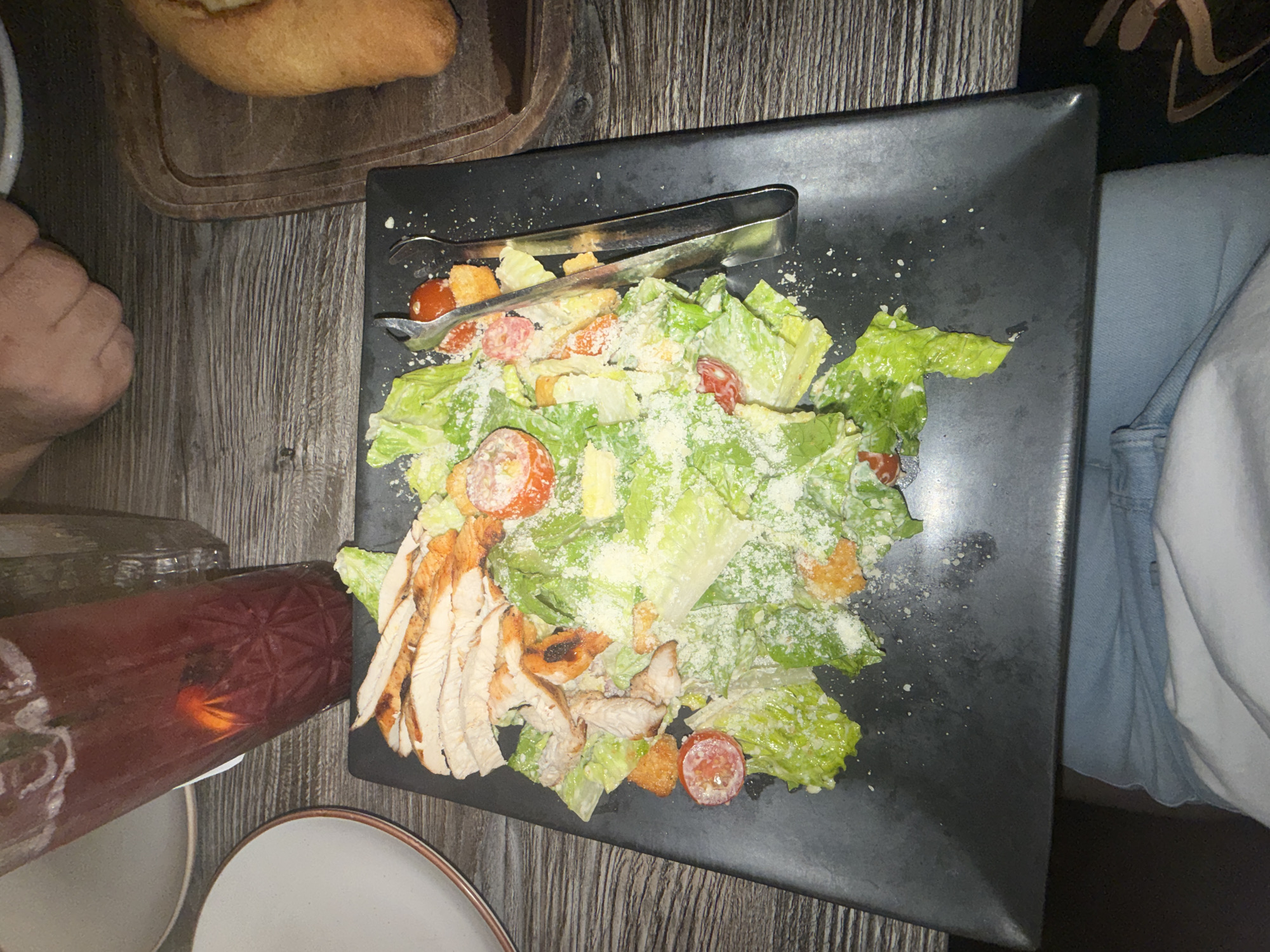 A top-down shot shows a dark-colored rectangular plate resting on a wooden-textured surface. The plate holds a salad featuring romaine lettuce, cherry tomatoes, croutons, sliced grilled chicken, and a sprinkle of shredded cheese. A pair of salad tongs rests on top of the salad.
To the left of the plate, there is a glass filled with a dark red liquid, along with a white plate. Above the plate, a wooden board with a bread item rests. To the right, a person wearing light blue jeans is partially visible.
The image was likely taken indoors at a restaurant. Further information is needed to confirm if it is located in Glenview, United States of America.