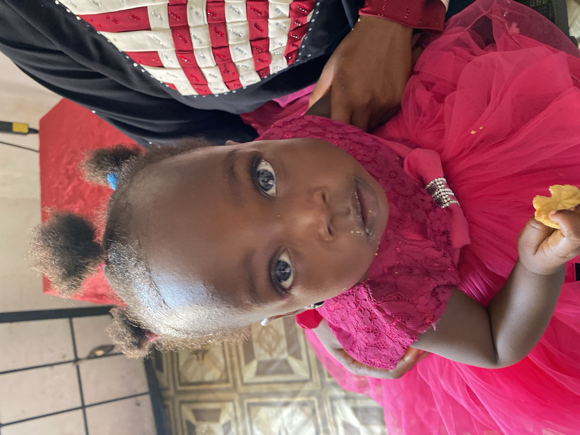 A close-up, rotated image captures a young child held by an adult indoors. The child, with a dark complexion and dark, expressive eyes, looks directly at the camera. Their hair is styled in two small afro puffs on the sides of their head. They are dressed in a vibrant fuchsia pink dress, featuring a lace bodice and a voluminous, layered tulle skirt. A silver, beaded embellishment adorns the dress near the child's left arm. Small crumbs or specks of food are visible on the child's face, around the mouth and nose. In their right hand, the child holds a light-colored, possibly baked, item like a biscuit or cookie.
Only portions of the adult holding the child are visible. The adult wears a dark garment, likely black, with a distinctive red and white checkered or woven pattern on the upper body, embellished with small reflective elements or sequins. A dark-skinned arm and hand of the adult are seen, presumably supporting the child.
The background indicates an indoor setting. A solid red surface, possibly a wall or table, is partially visible behind the child's head. The floor features square tiles with a geometric pattern, appearing in shades of cream and brown. The lighting is bright, suggesting it is daytime. No text is visible in the image.