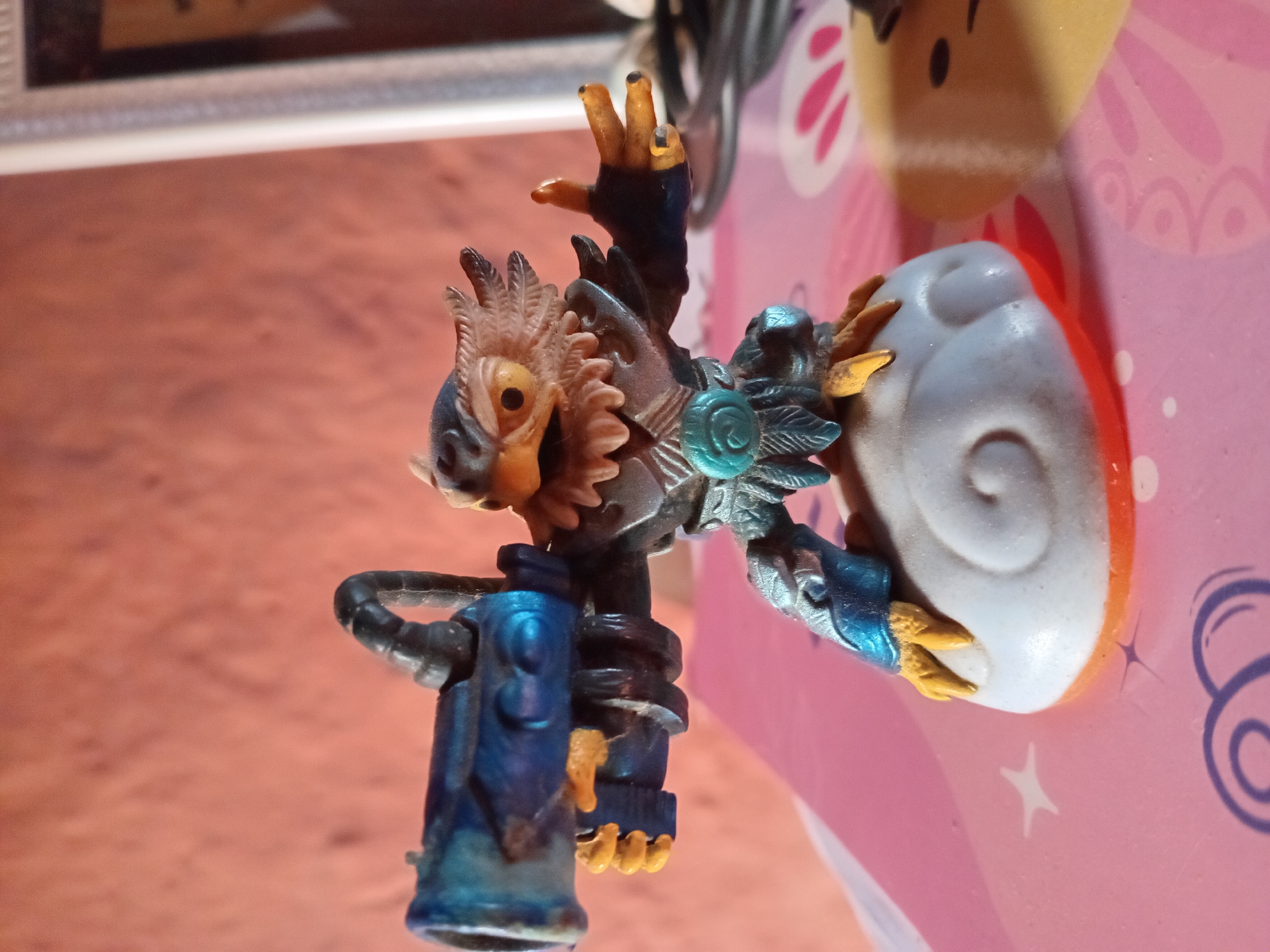 A plastic figurine of an owl-like creature with a large blue cannon is positioned on a white, cloud-shaped base. The creature has brown and tan feathers, yellow eyes, and talons. It is wearing metallic armor and a blue, circular emblem on its chest. The figurine is placed on a pink surface with white star and swirl patterns, suggesting it is on a table or desk. In the background, a textured coral-colored wall is visible, with a portion of a white picture frame to the right. The image appears to have been taken indoors, possibly in a home setting. The location of Juan Griego, Venezuela is not discernible from the provided image.
